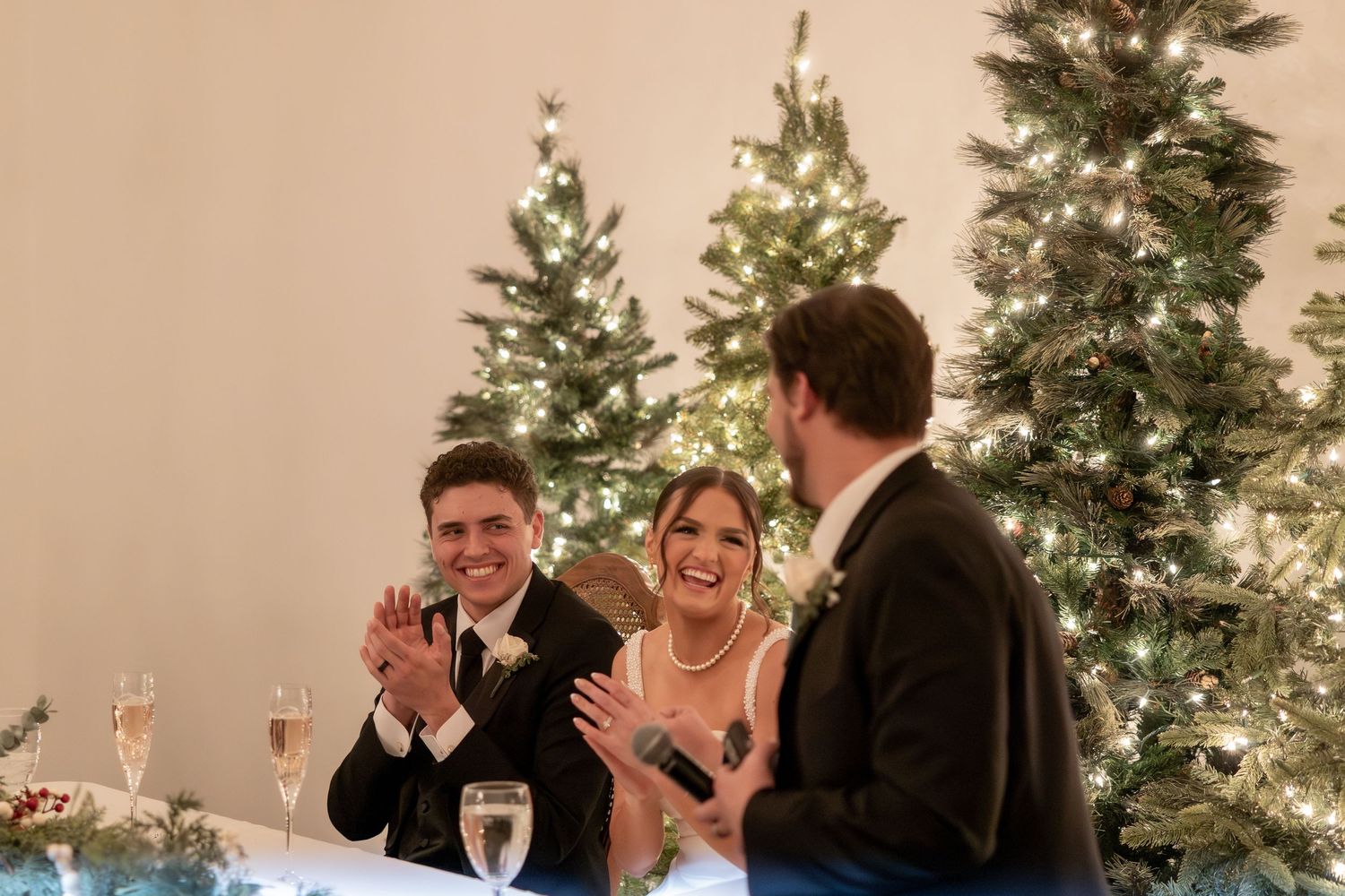 Bride and groom clapping and smiling as a guest speaks at their winter wedding reception with Christmas trees.