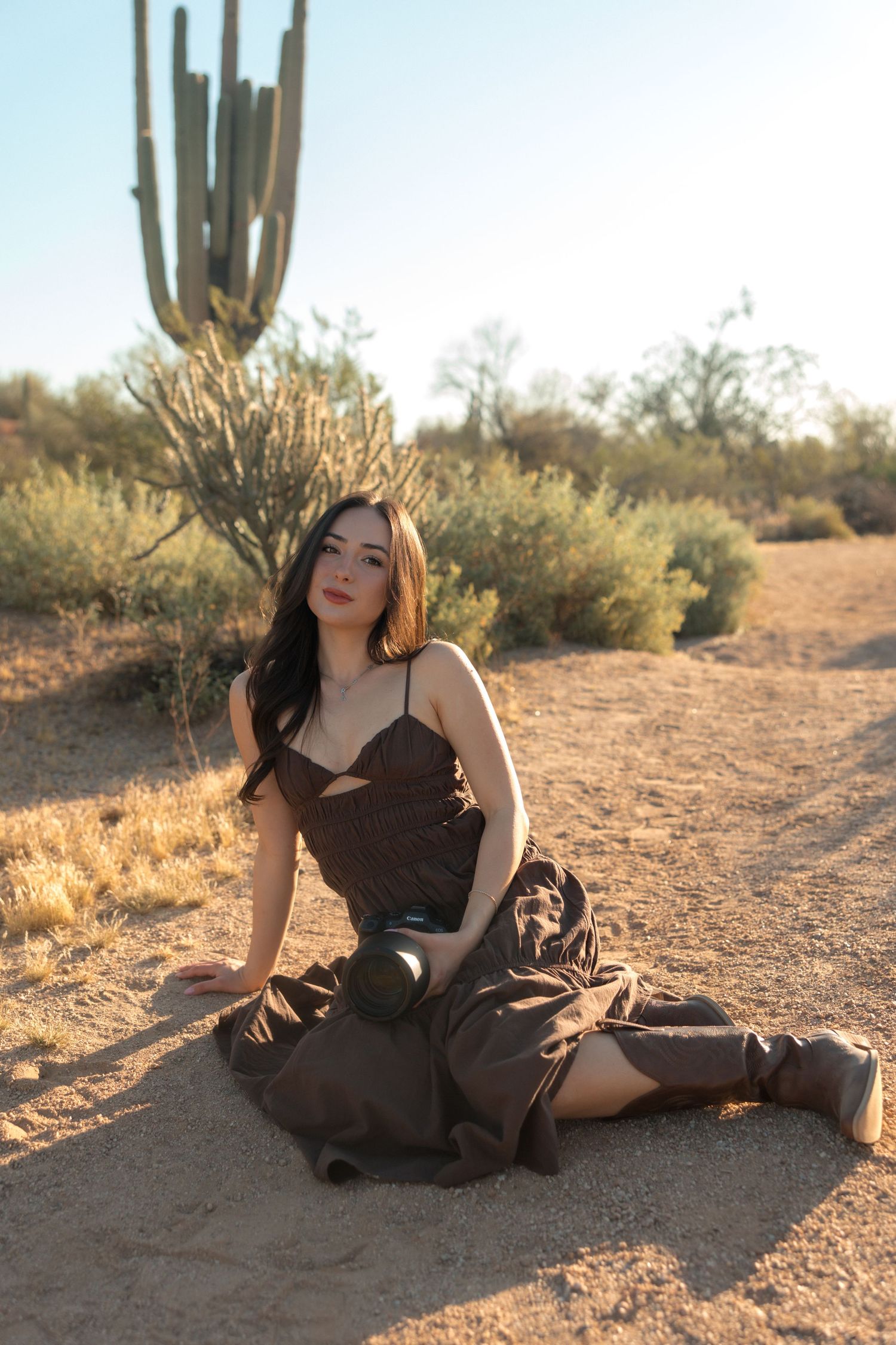 Young woman in brown sundress sitting on desert ground with saguaro cactus and blue sky behind her.