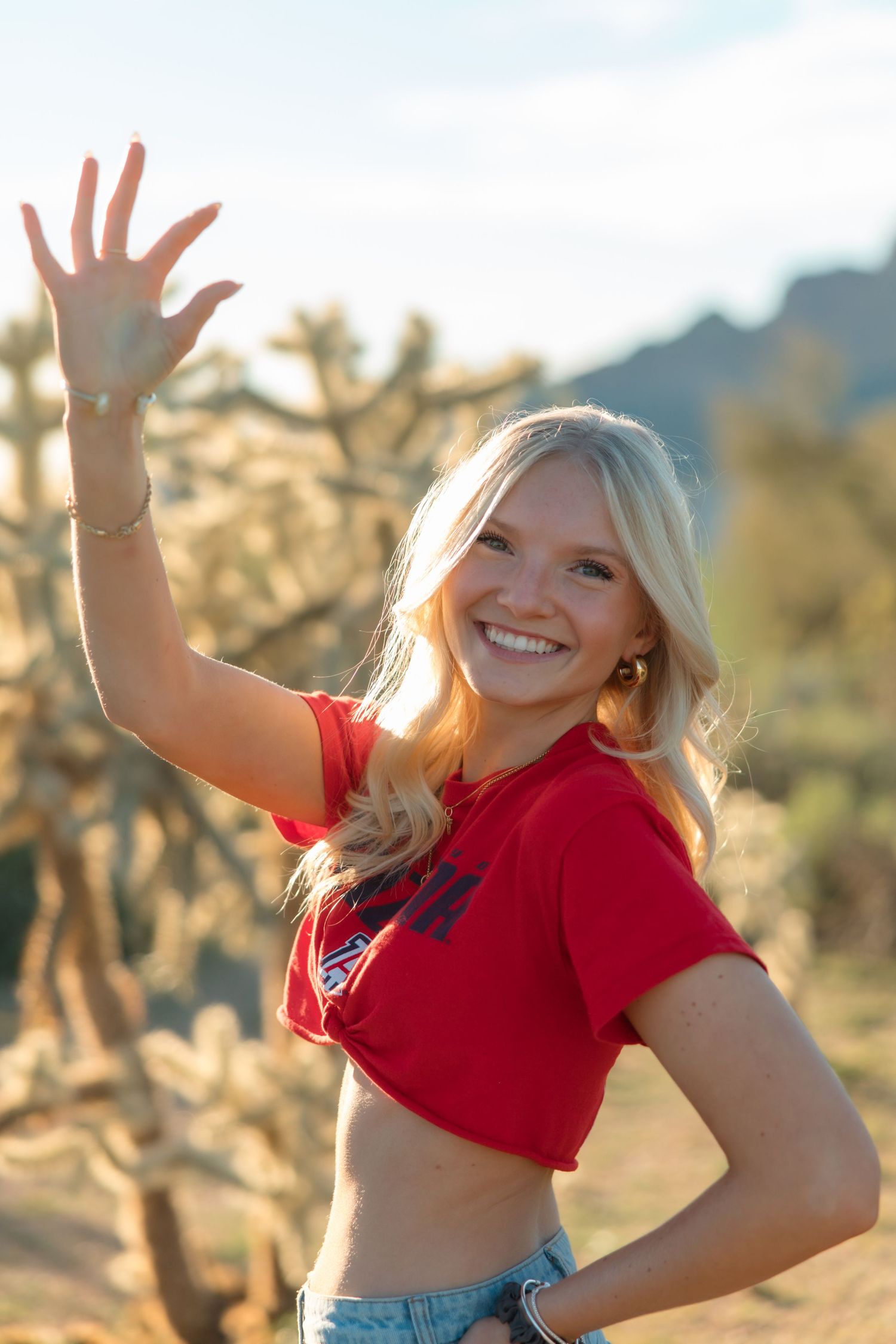 Smiling blonde woman in red crop top waving outdoors in desert landscape with cacti and mountains.