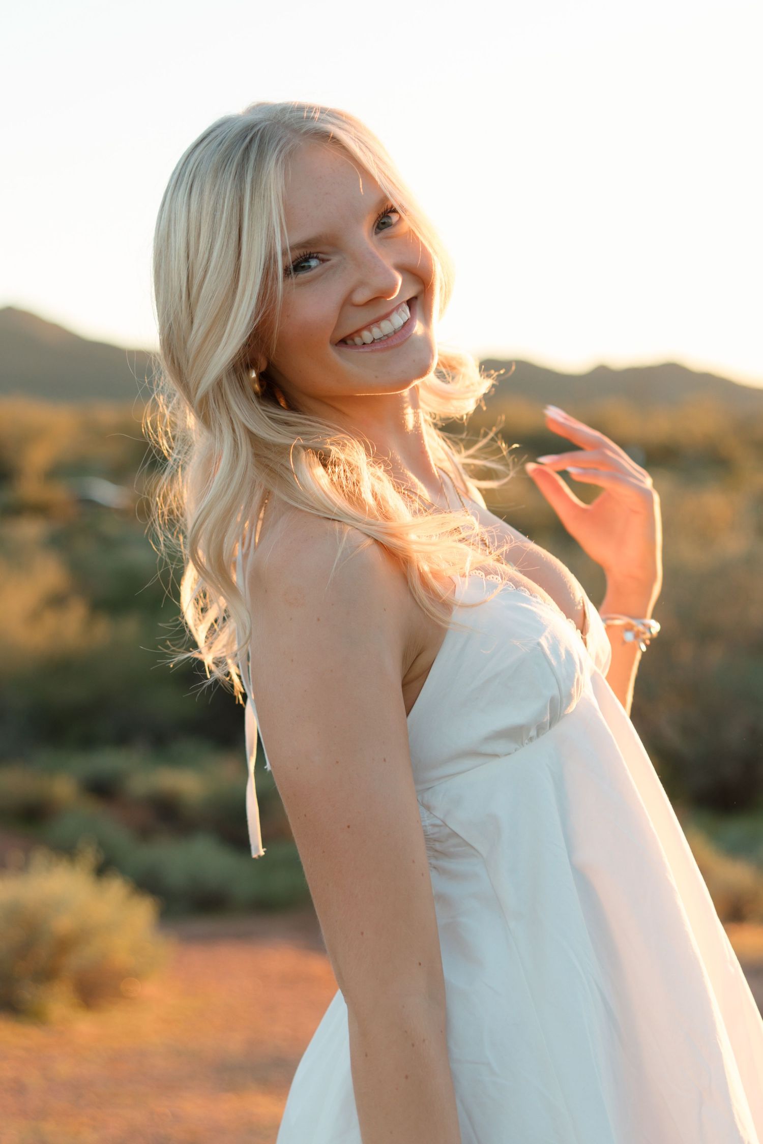 Smiling blonde woman in white dress poses outdoors at golden hour with desert landscape and mountains in background.