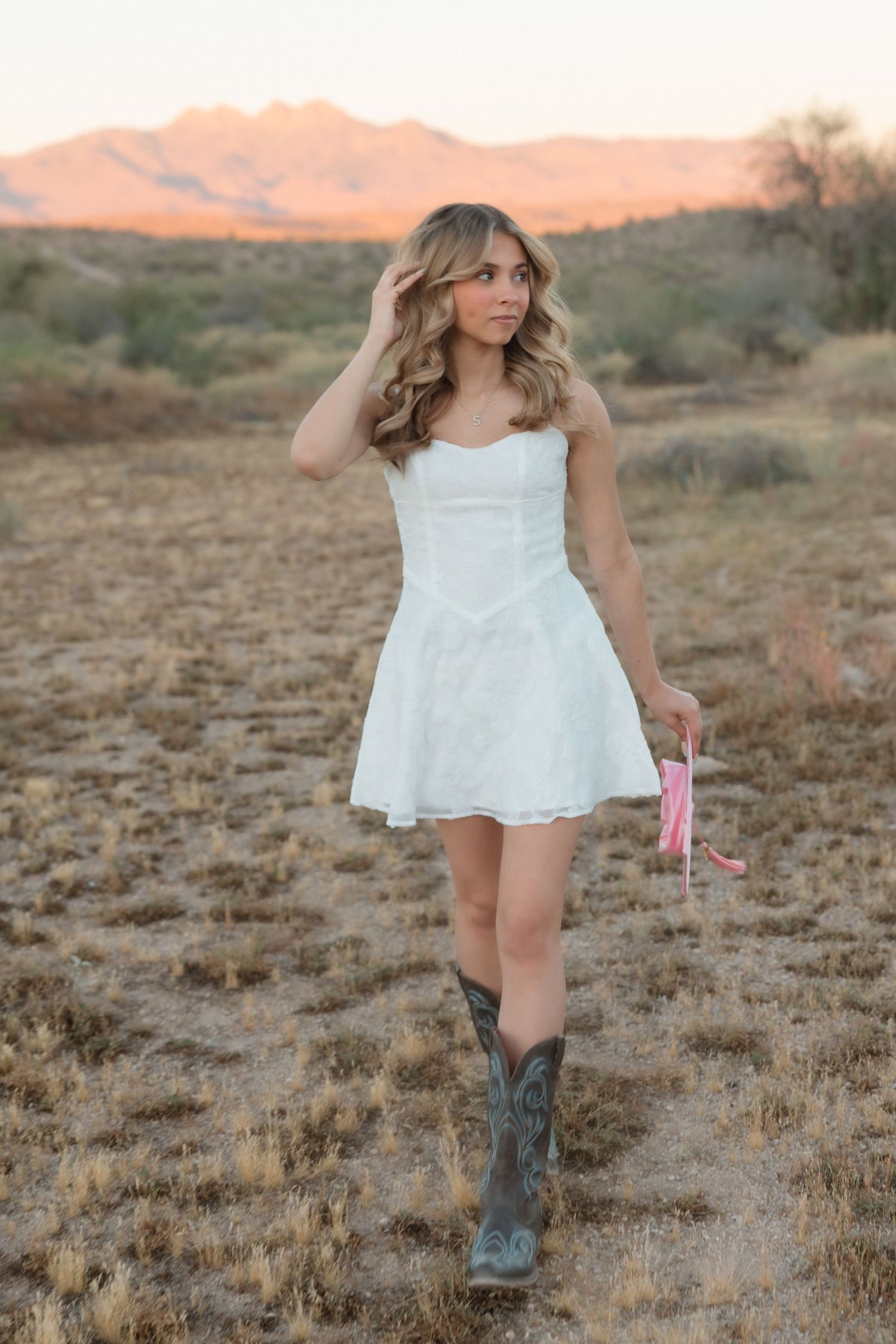 Young woman in white mini dress and cowboy boots holds pink ribbon in desert landscape at golden hour sunset.