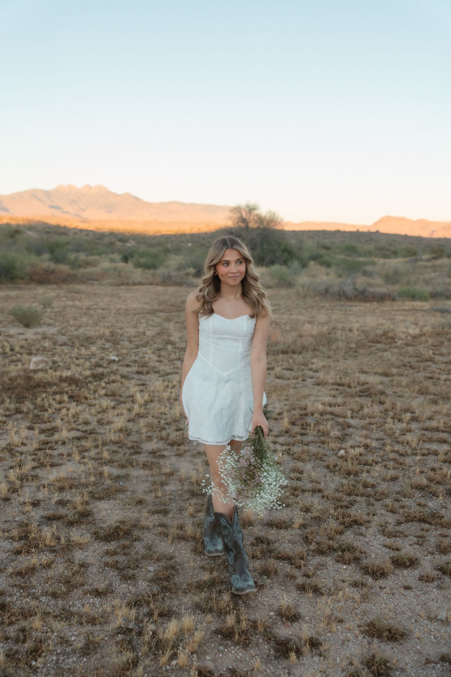 Young woman in white dress and cowboy boots holds wildflowers in a desert landscape at golden hour.