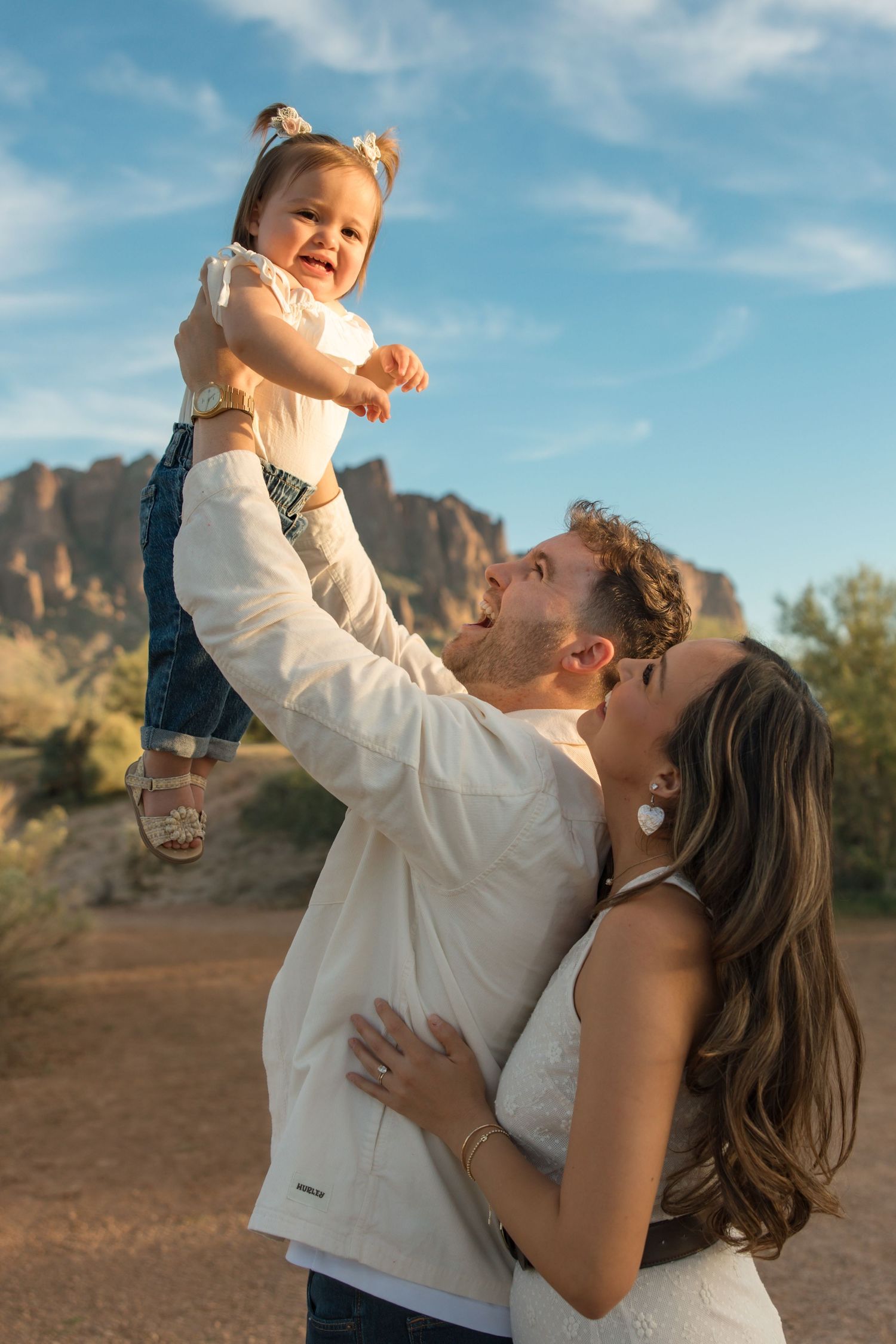 A couple lifts their laughing baby girl in a desert landscape with rocky mountains and blue sky behind them.