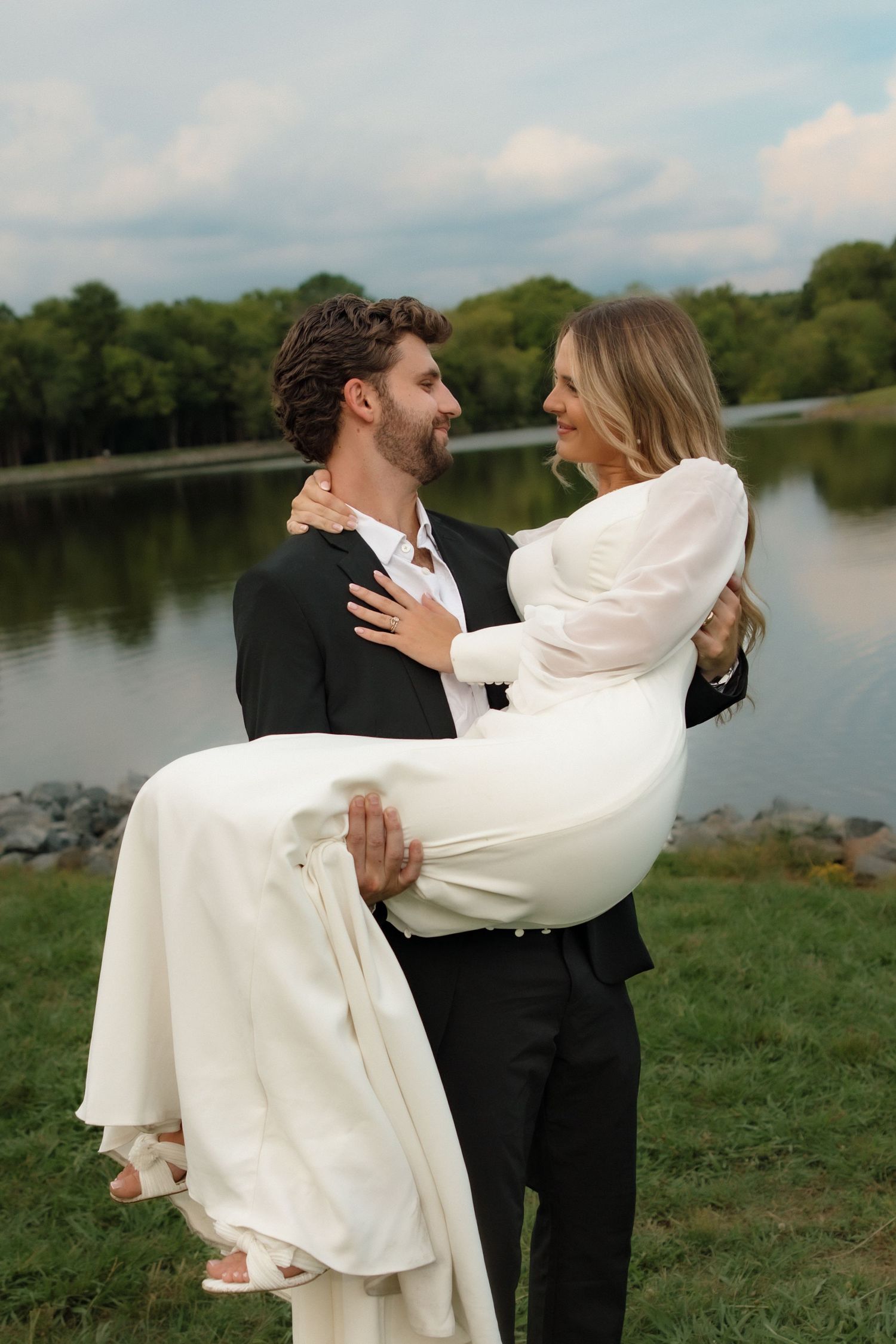 Groom in black suit carrying bride in white dress near a serene lake surrounded by lush greenery.