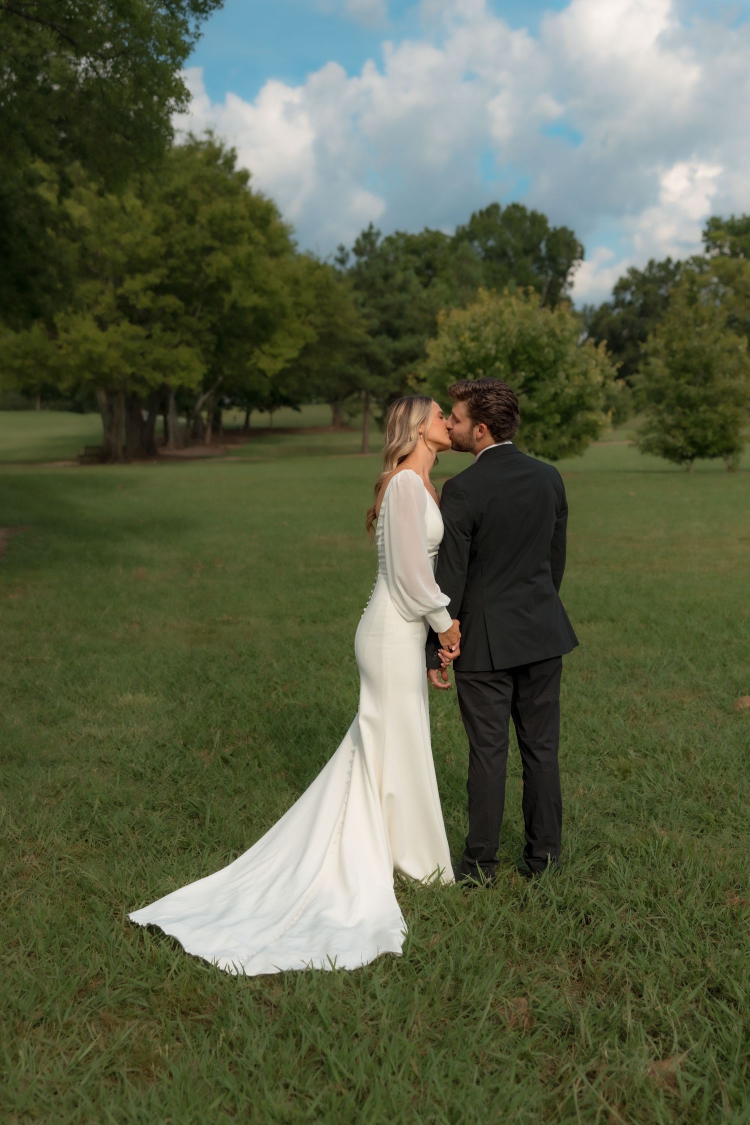 Bride and groom sharing a romantic kiss in a lush green park, bride wearing elegant white gown with long train.