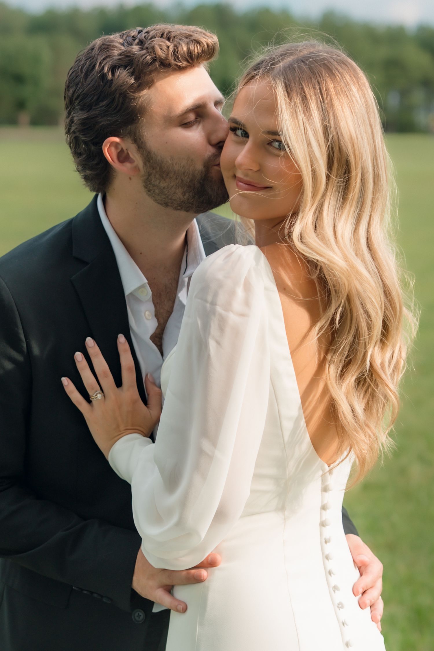 A groom in a black suit kisses his bride on the cheek as she smiles, wearing a white long-sleeve wedding dress outdoors.