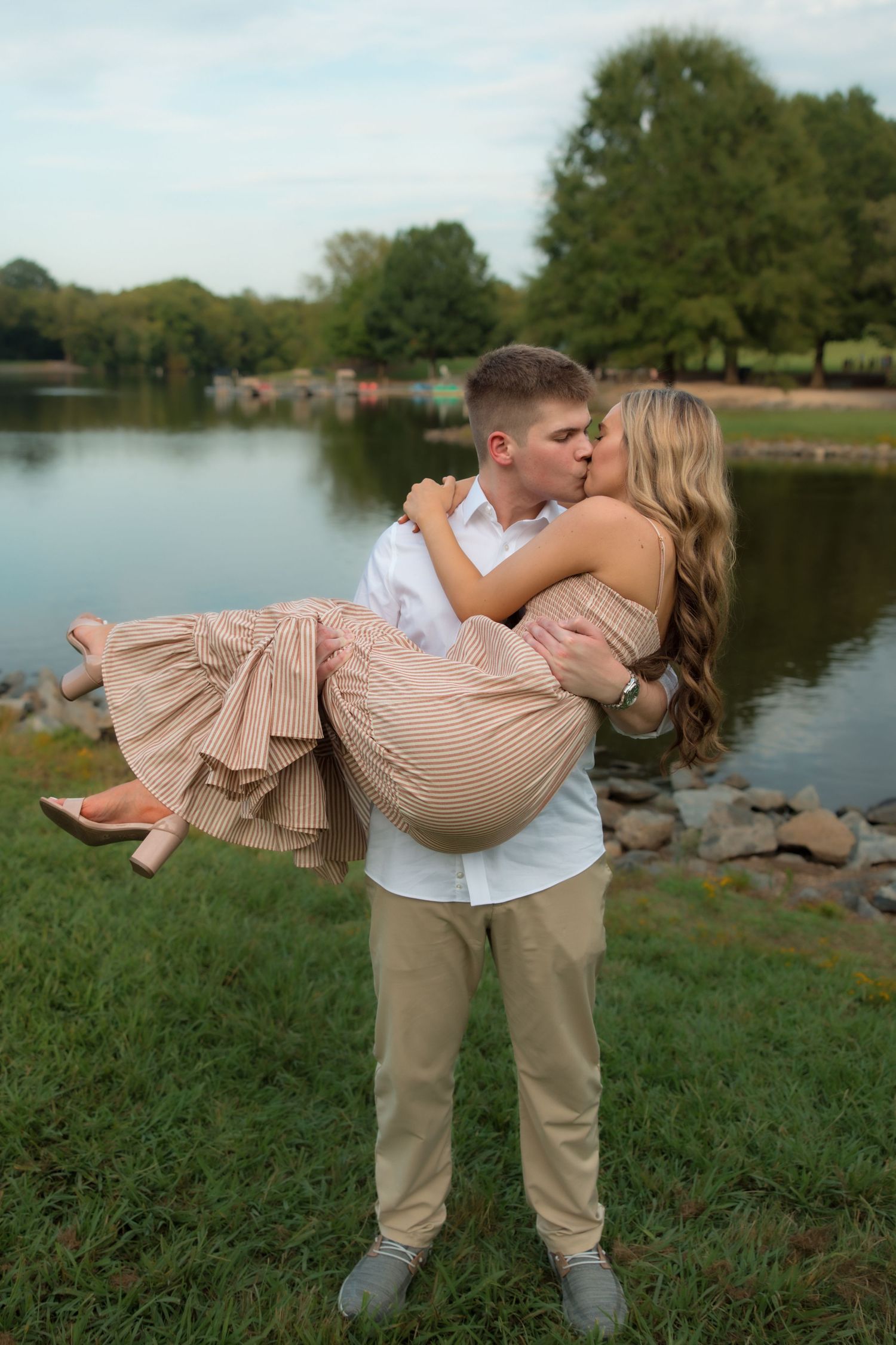 A couple shares a romantic kiss as the man lifts the woman in his arms near a serene lake in a park.