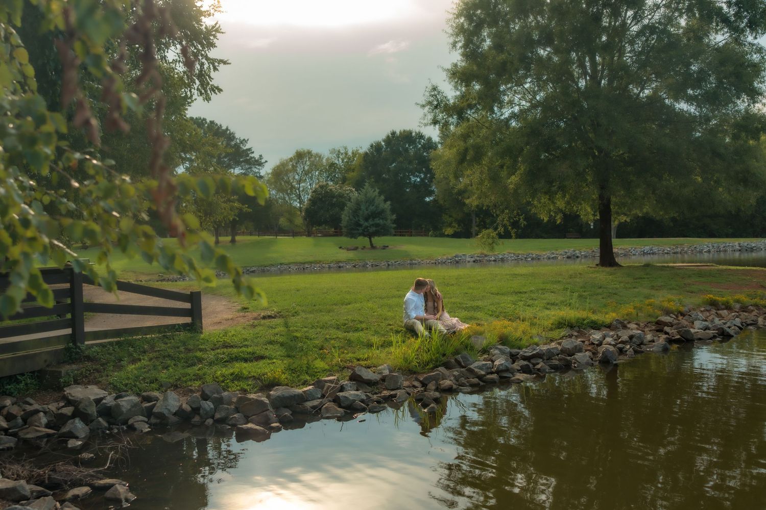 Couple sitting by a serene pond at golden hour in a lush green park with trees and a wooden fence.