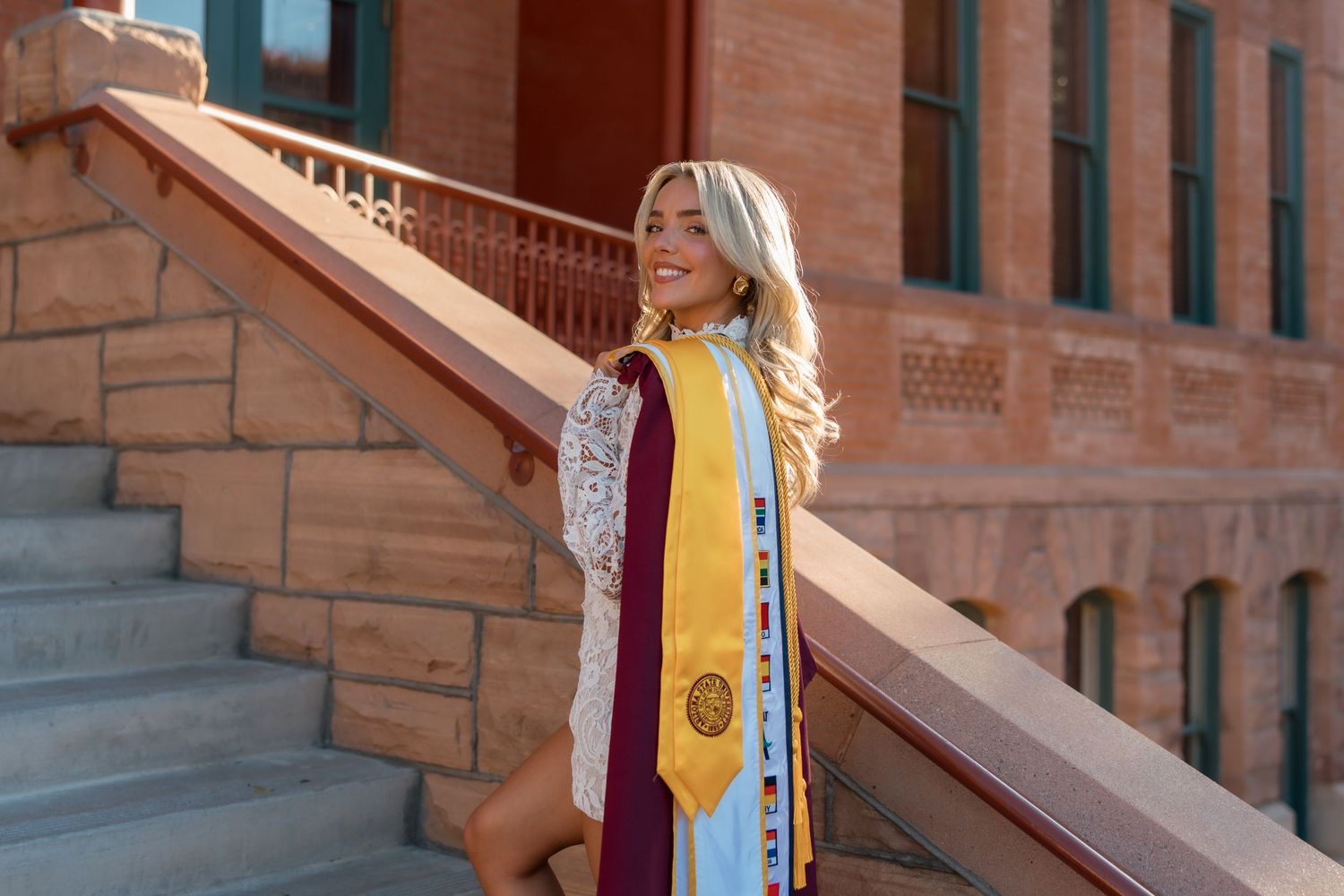 Blonde female graduate smiling on brick staircase wearing maroon gown and gold ASU honor stole.