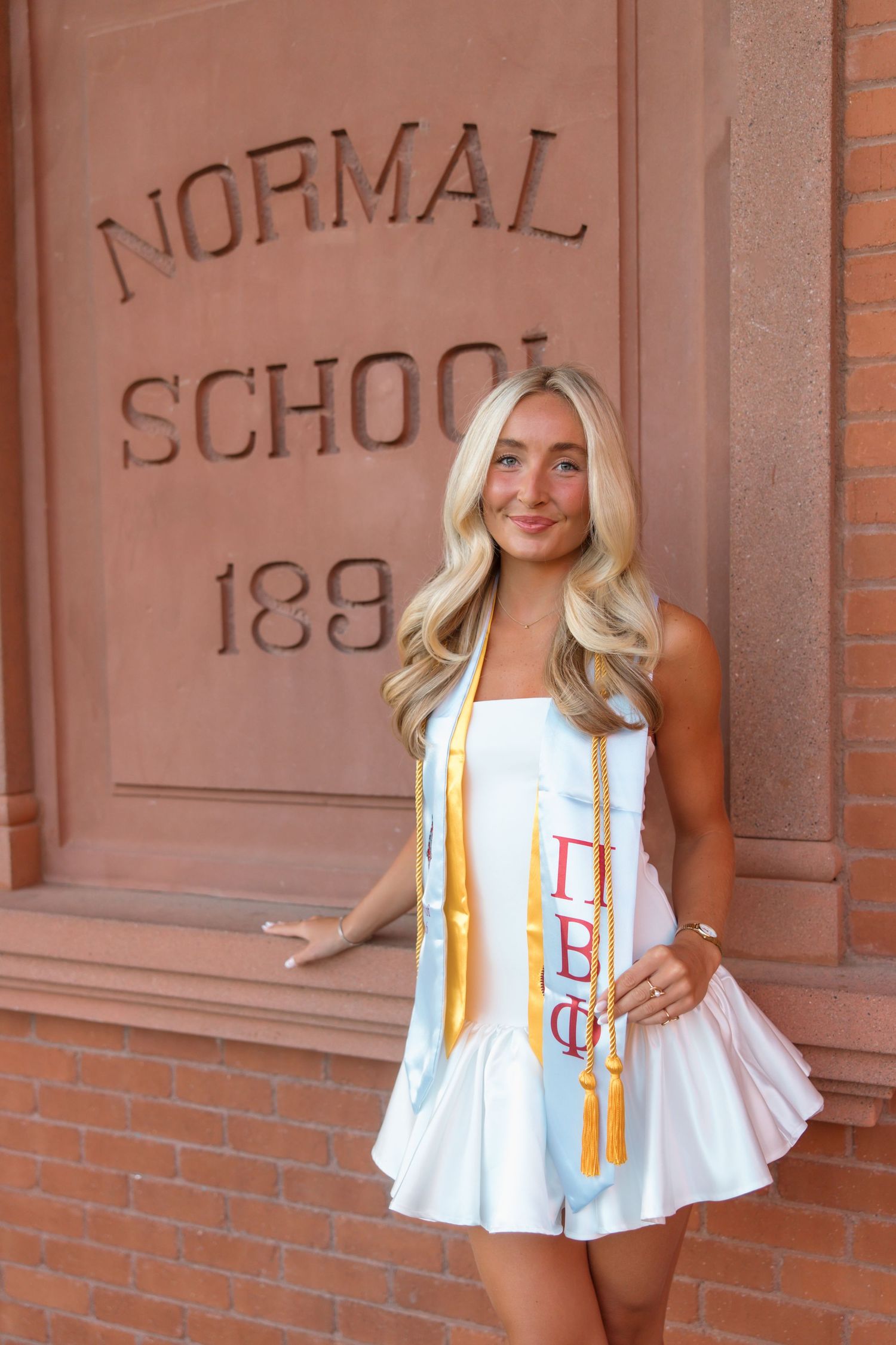 Blonde woman in white dress with Pi Beta Phi stole posing by Normal School 1889 brick building.