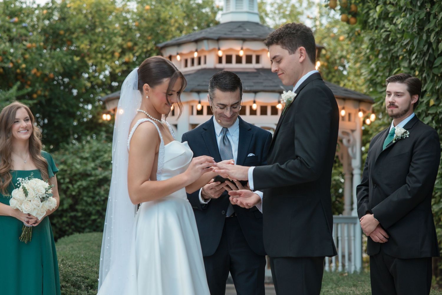 Groom places wedding ring on brides finger during outdoor gazebo ceremony with wedding party watching.