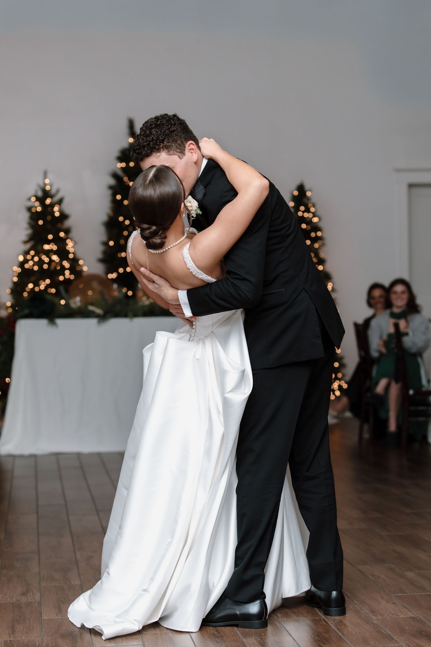 Bride and groom share their first dance, embracing closely in a romantic wedding reception with Christmas trees.