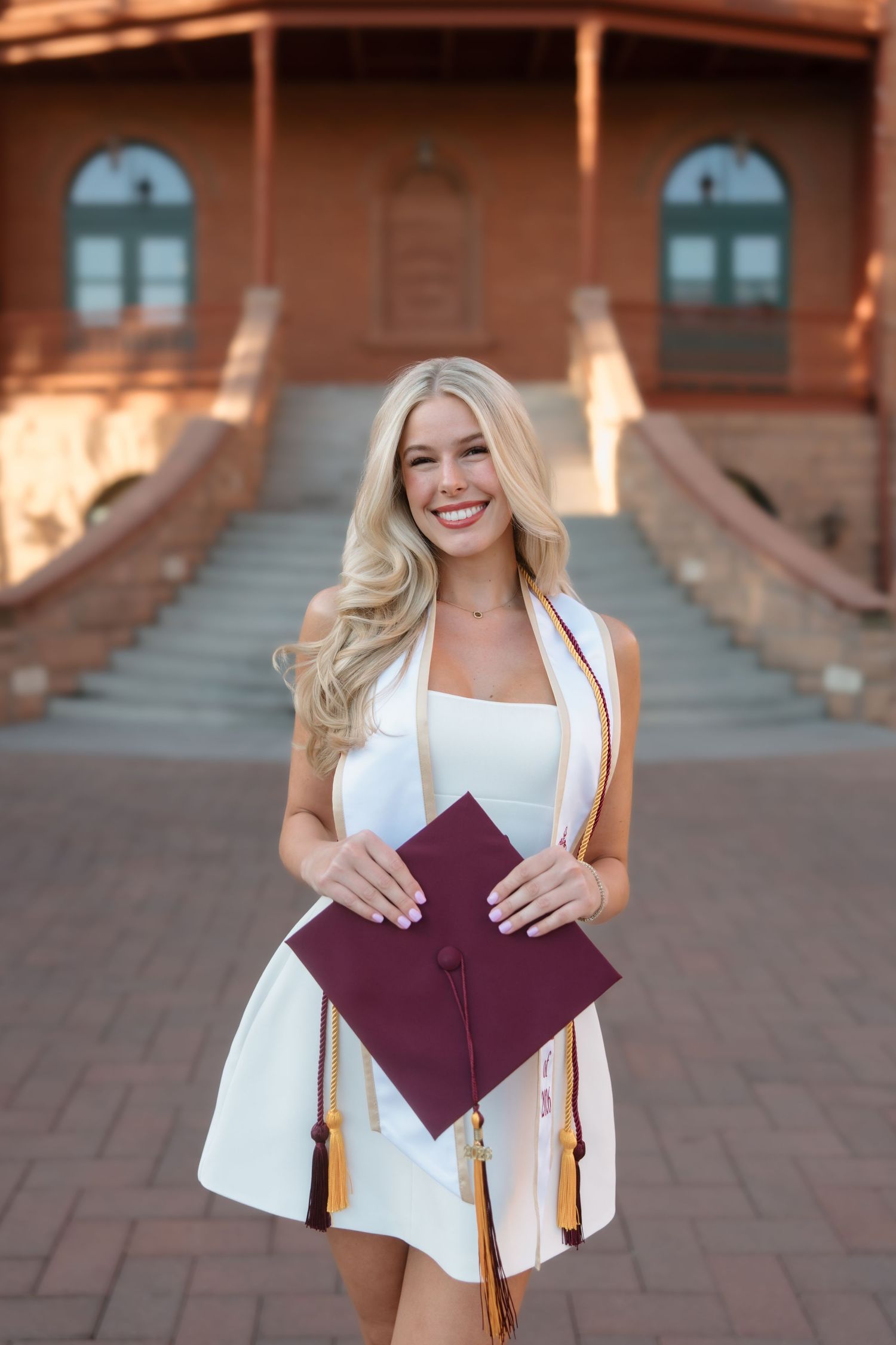 Smiling blonde graduate holds maroon cap wearing white dress with gold and maroon honor stoles outside ASU building.