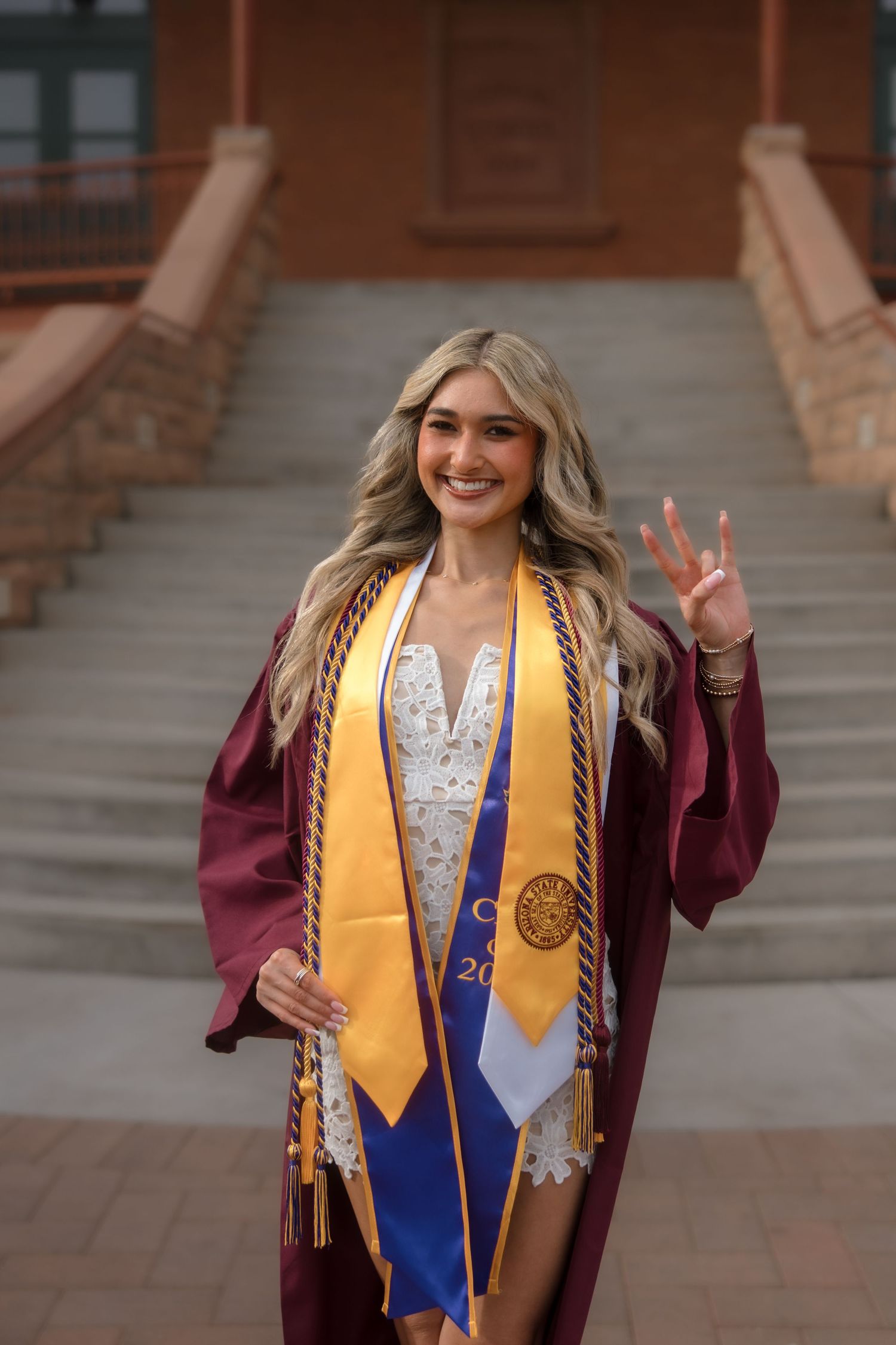 Smiling ASU graduate in maroon robe and gold honor stole giving pitchfork hand sign on historic campus staircase.