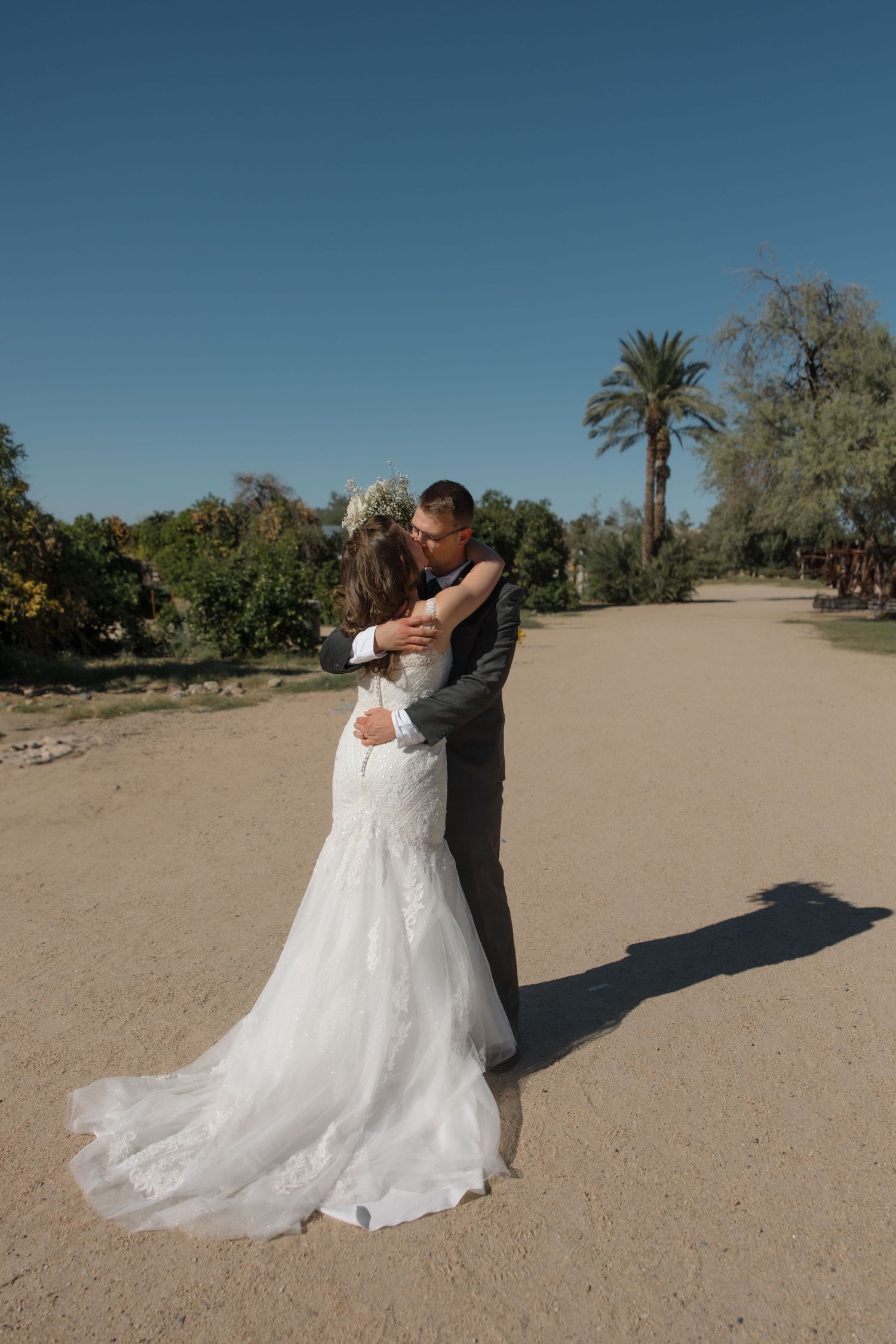 Bride and groom share romantic embrace on sandy path surrounded by palm trees and lush greenery outdoors.