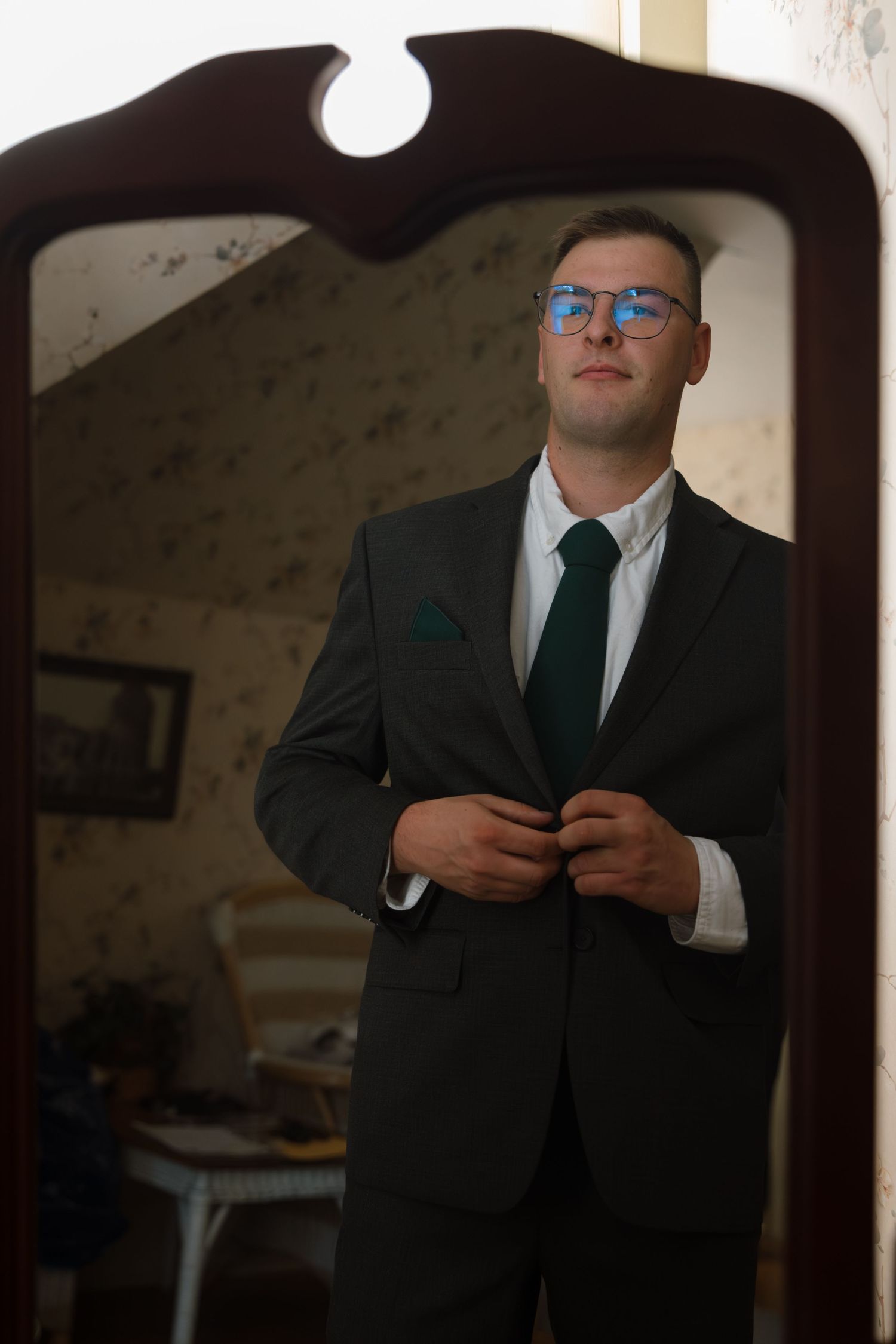 Man in dark suit, green tie, and glasses buttoning jacket while standing in front of ornate mirror.