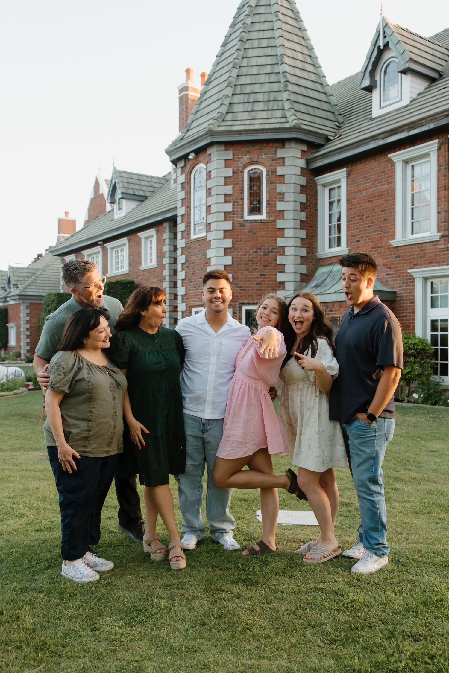 A joyful family group poses together on a green lawn in front of a large brick Tudor-style home at dusk.