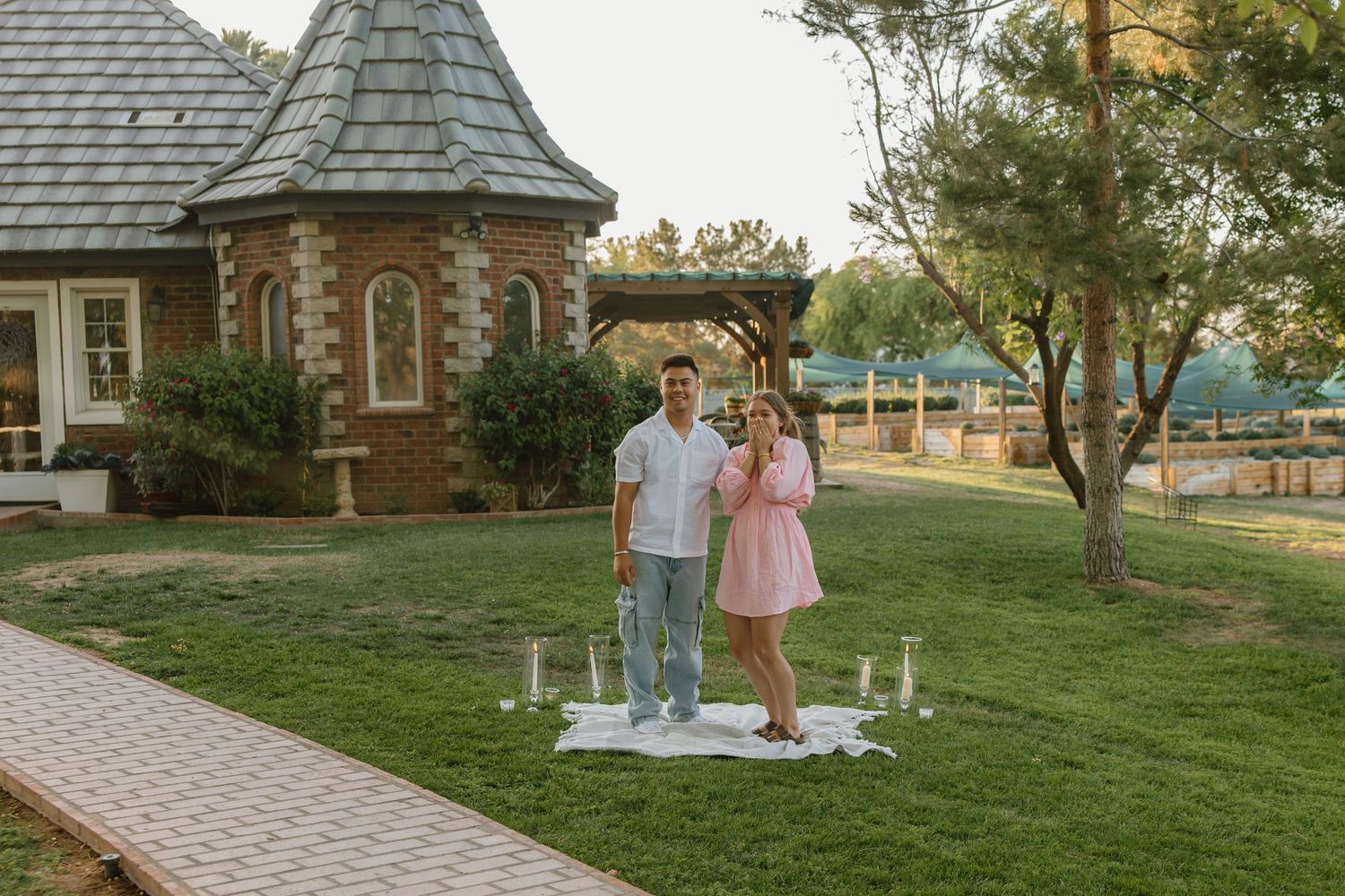Couple standing on a decorated lawn beside a charming brick chapel at golden hour.