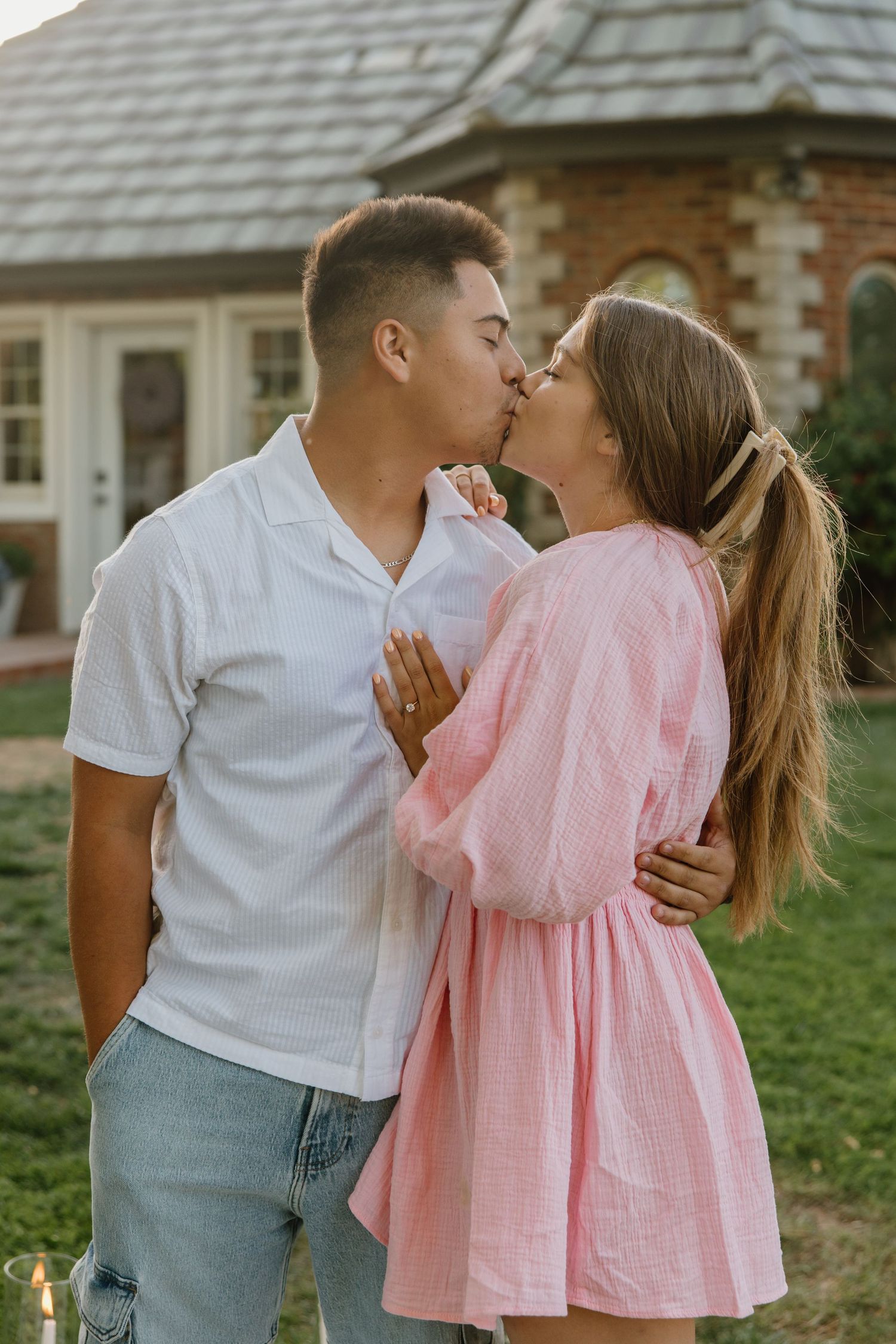 A young couple shares a romantic kiss outdoors, with the man in a white shirt and woman in a pink dress embracing.