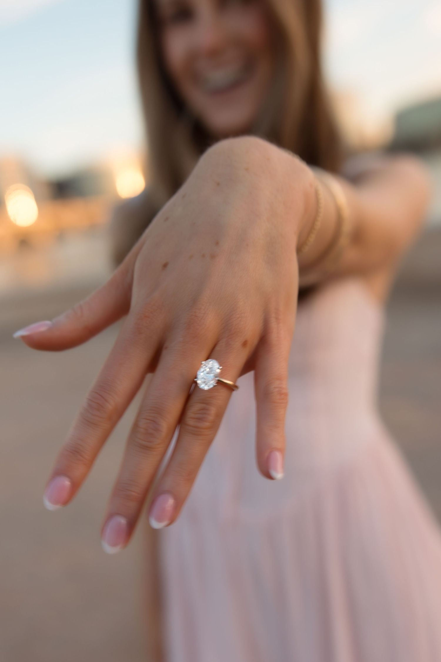 Close-up of woman showing oval diamond solitaire engagement ring on outstretched hand at sunset.