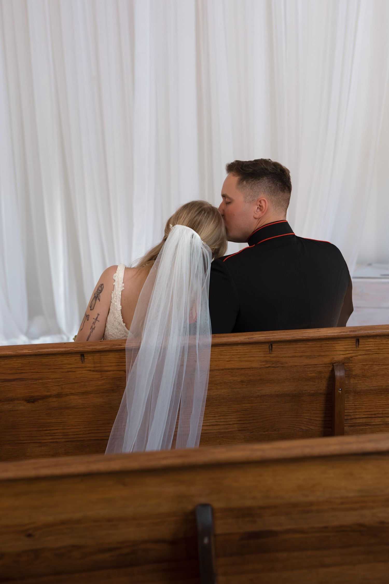 Bride and groom share a kiss on wooden church pew during wedding ceremony with white drape backdrop.