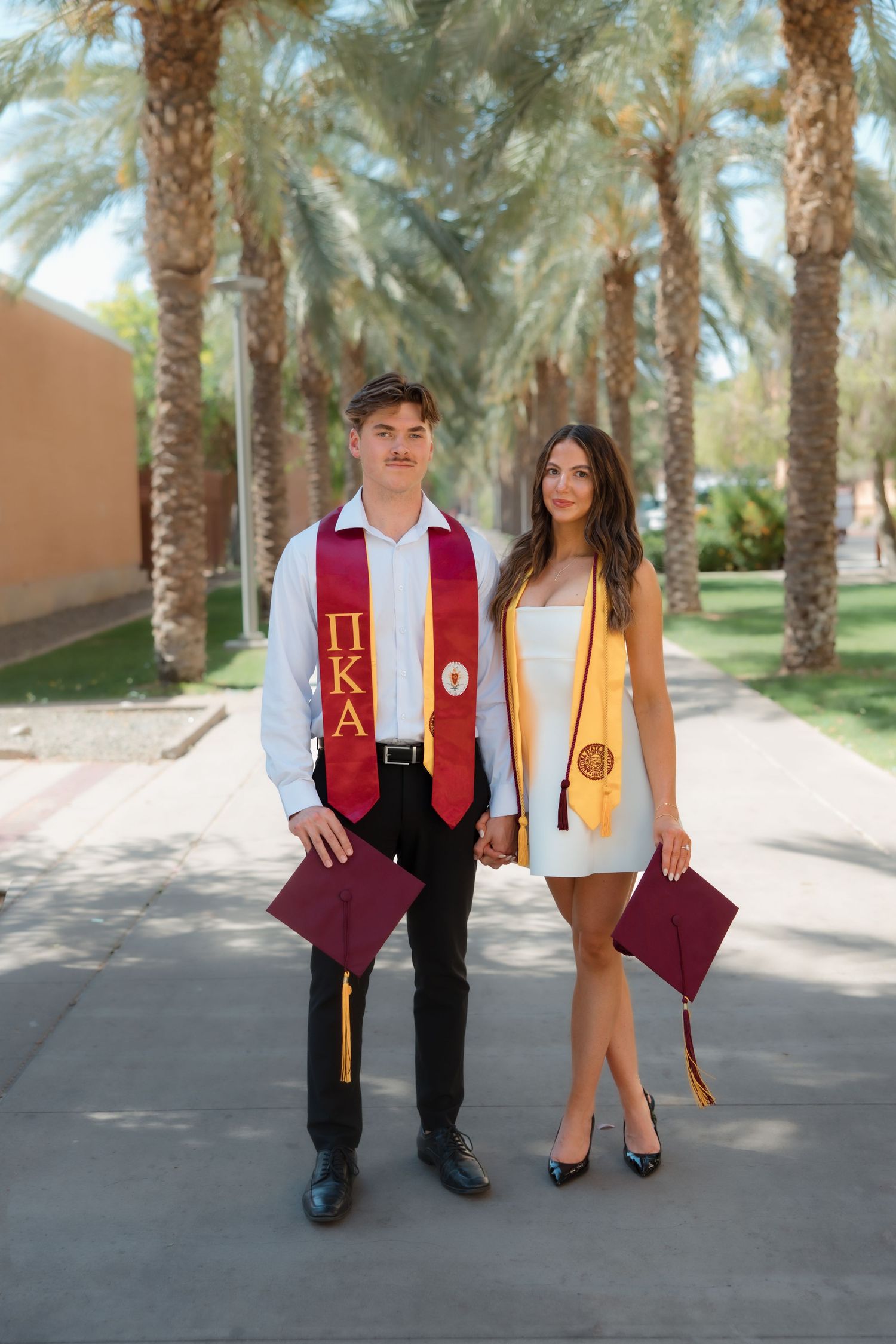 Graduating couple holding caps and holding hands on palm tree-lined campus walkway.