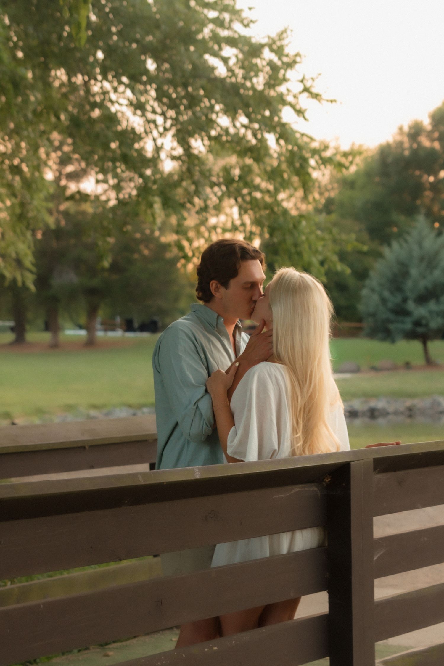 A young couple kisses intimately on a wooden bridge in a serene park during sunset.