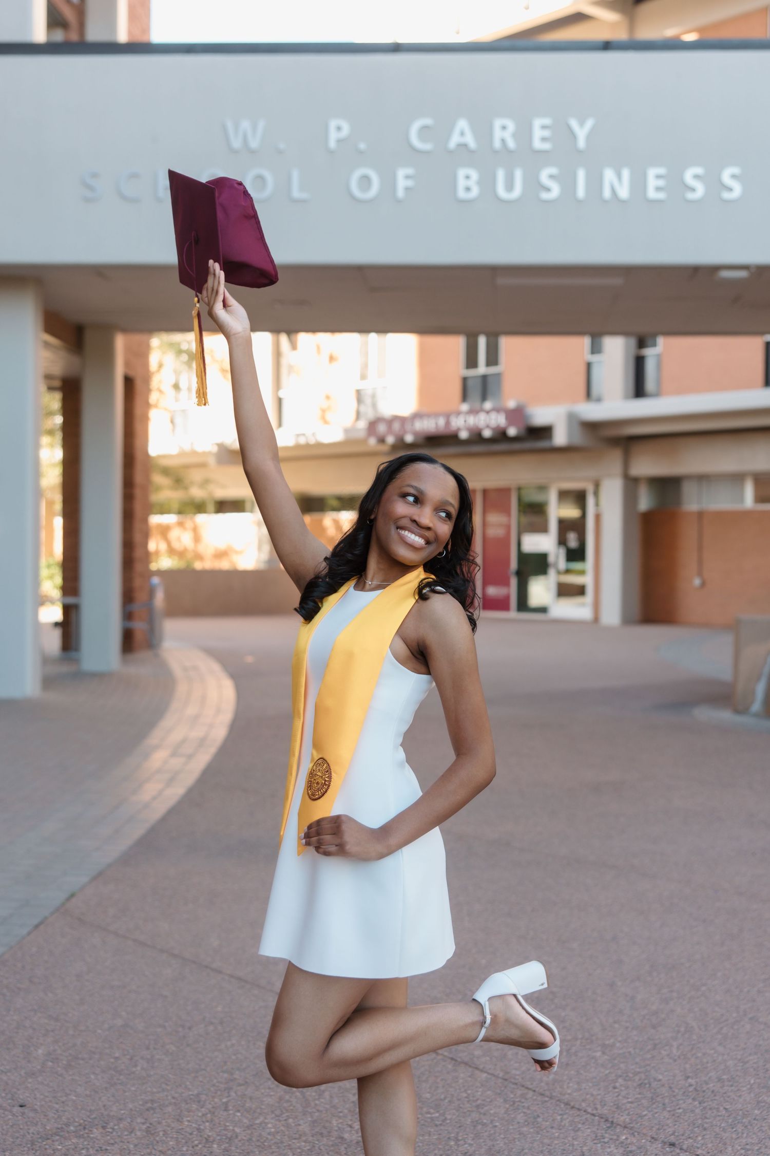 Smiling graduate in white dress raises cap in front of W.P. Carey School of Business building.