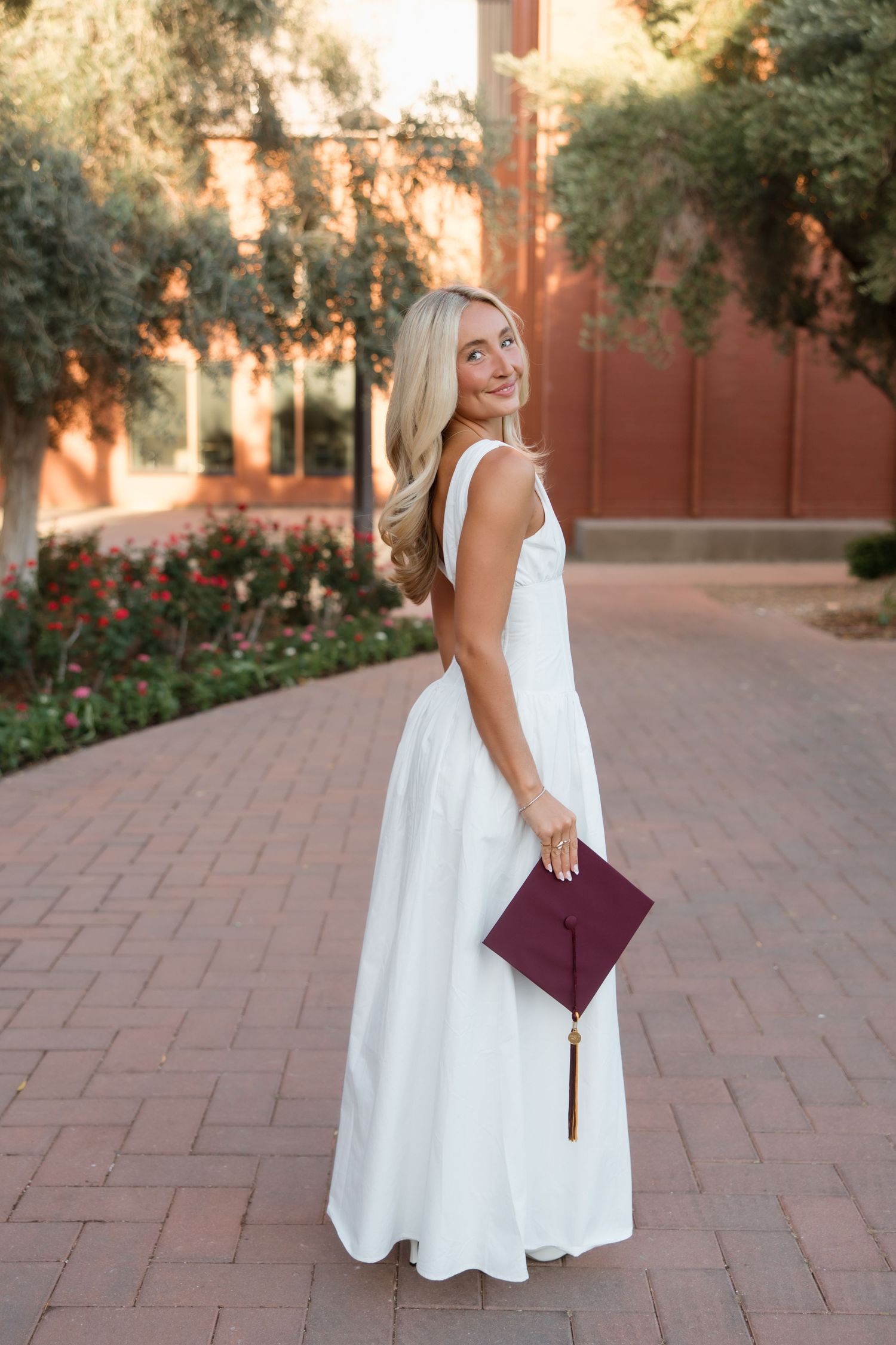 Smiling blonde woman in white gown holding burgundy graduation cap on brick path with red building backdrop.