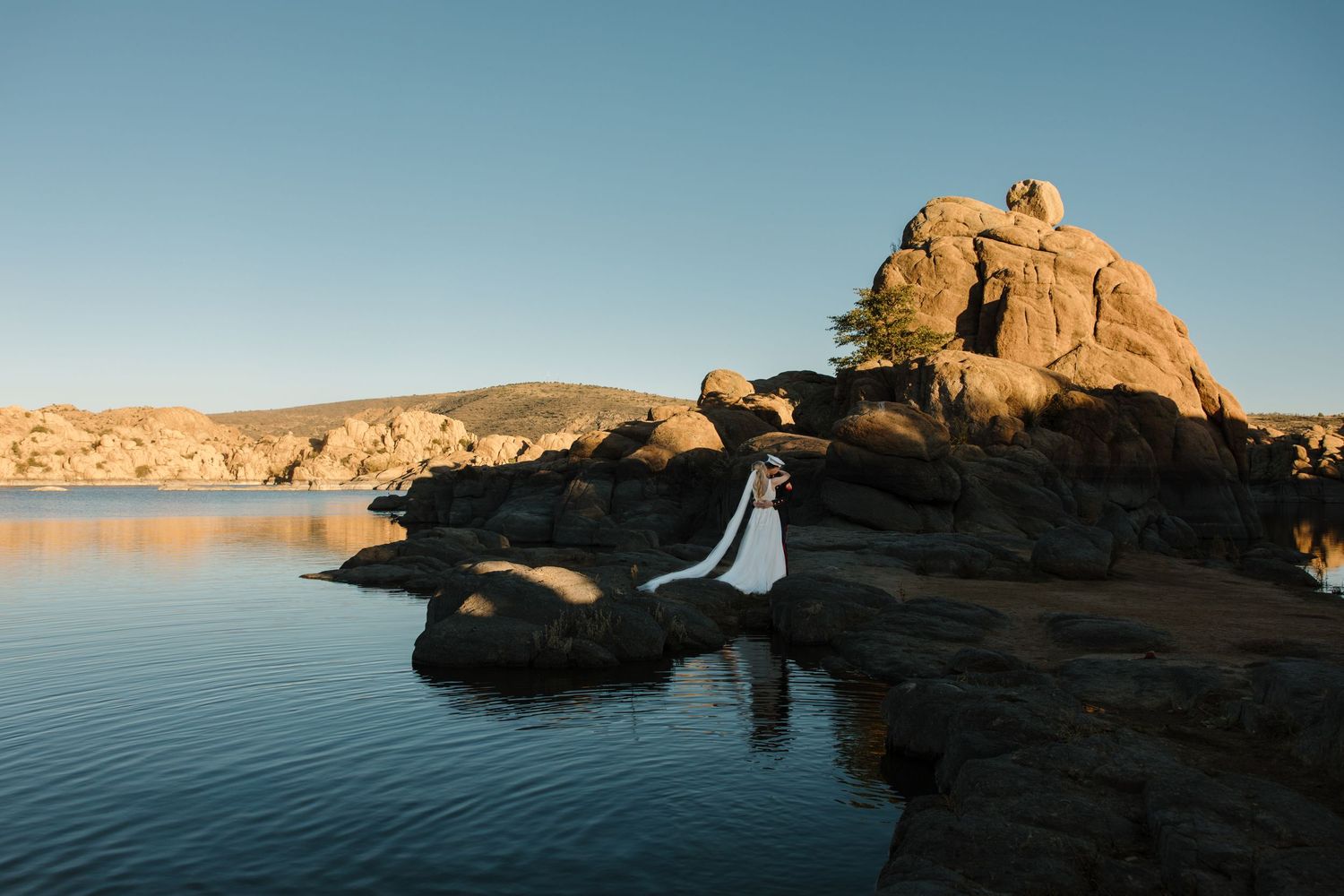 Bride and groom stand on rocky shore beside serene lake with dramatic boulder formations.