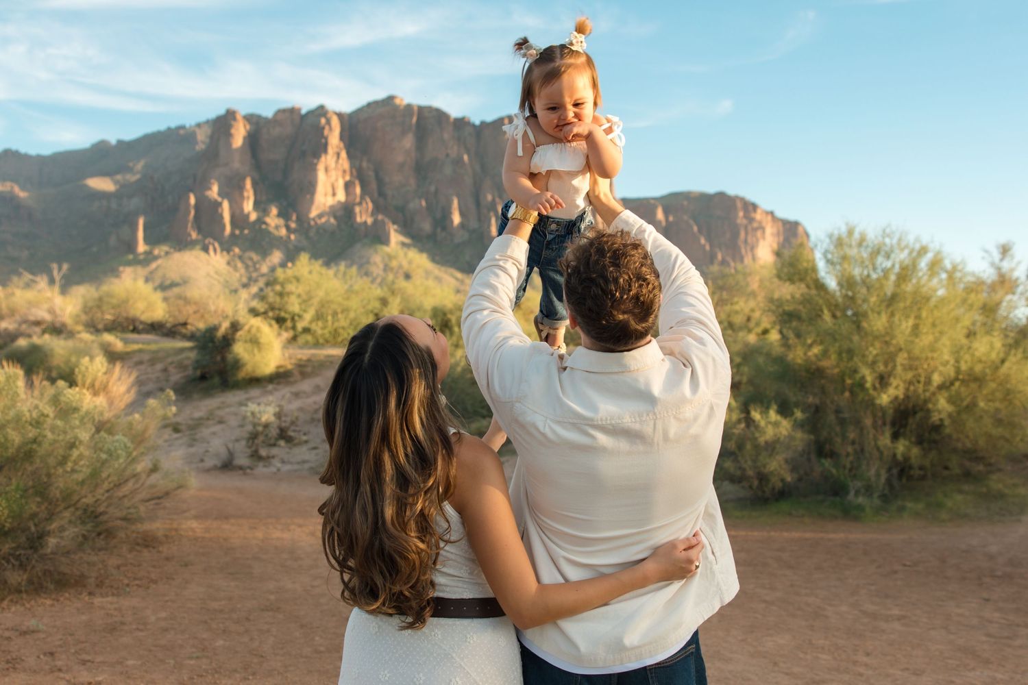 A family poses in the Arizona desert, father lifting toddler girl while mother embraces him, Superstition Mountains behind them.