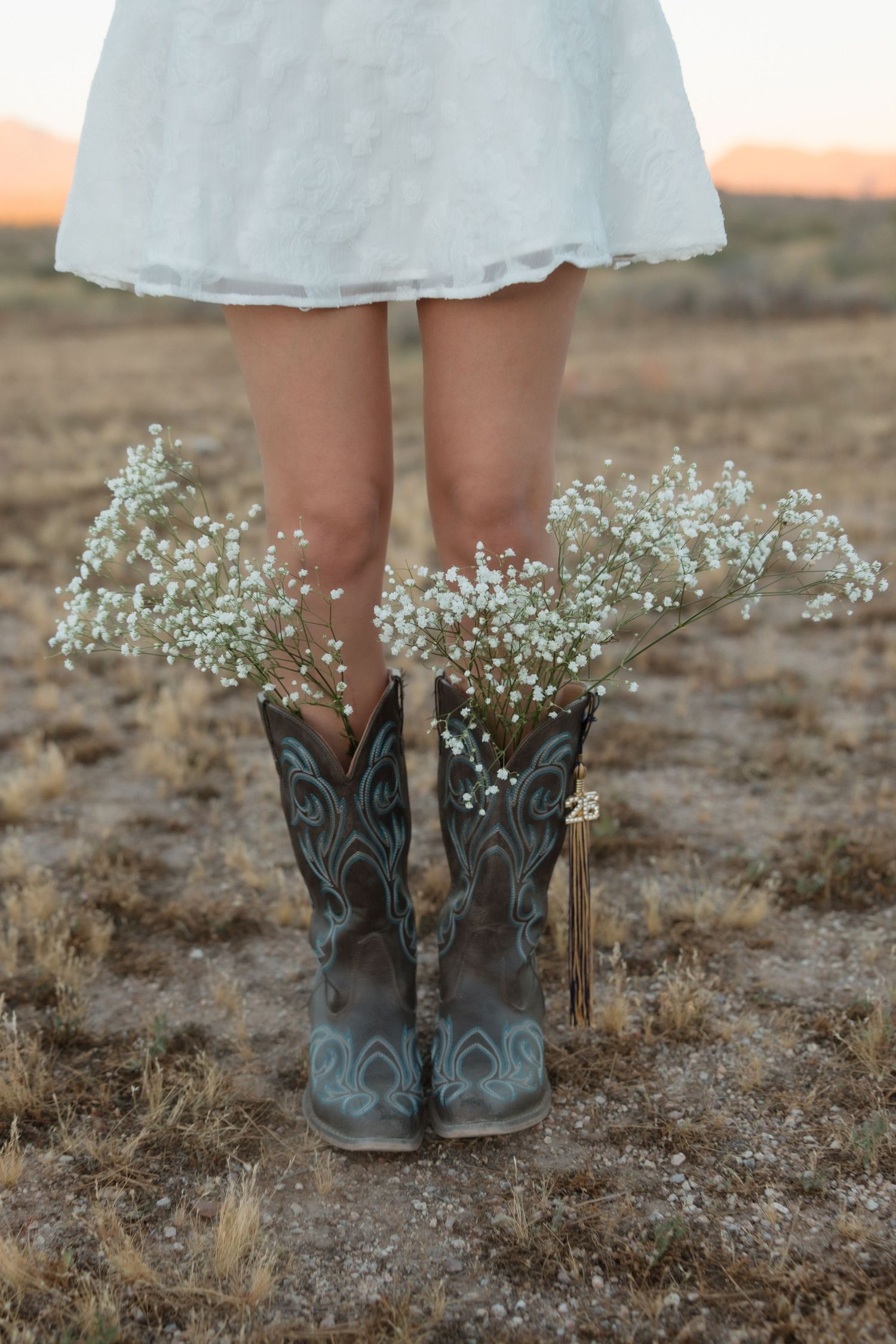 Woman in white dress wearing cowboy boots filled with baby's breath flowers standing in a rustic outdoor field at sunset.