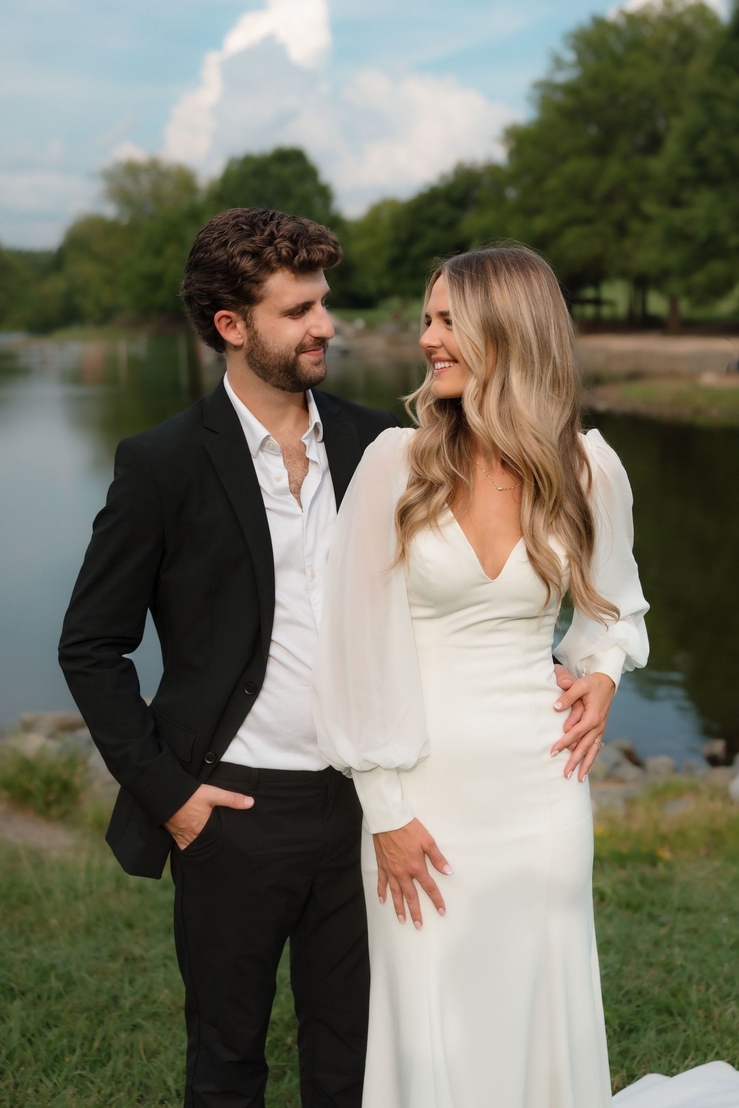 Elegant couple in black suit and white gown smiling together near a serene lakeside setting.