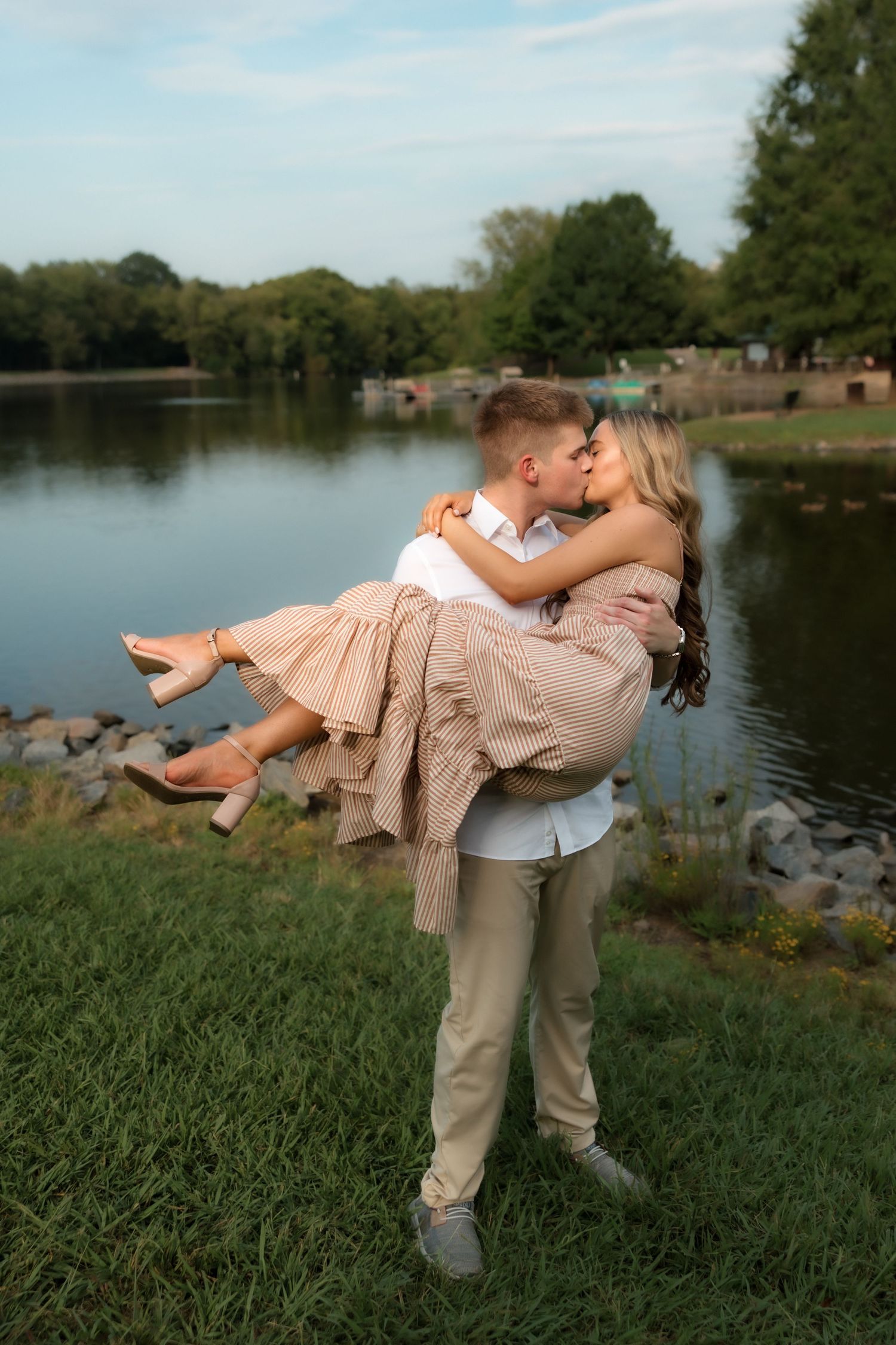 A couple shares a romantic kiss as the man lifts the woman in his arms by a serene lake in a lush park.