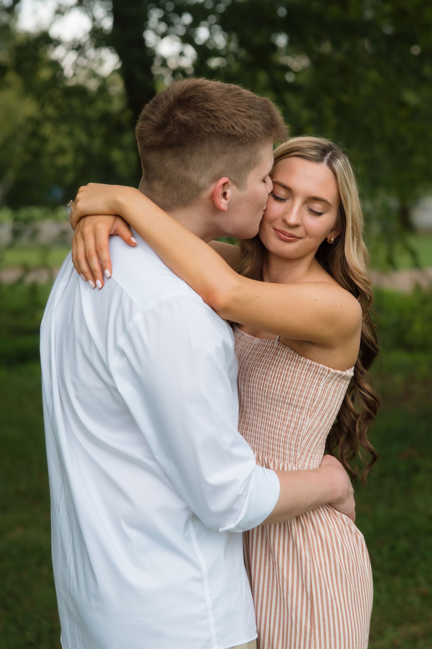Young couple embracing outdoors, woman in pink dress smiling with eyes closed as man in white shirt kisses her cheek.