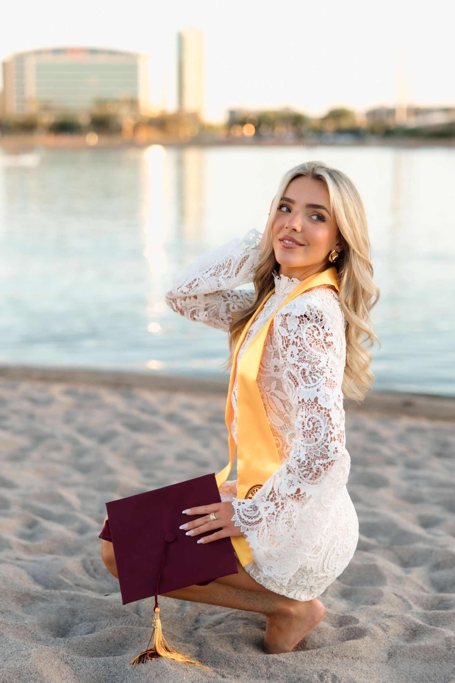 Blonde graduate in white lace dress with gold stole poses on sandy beach holding maroon cap.