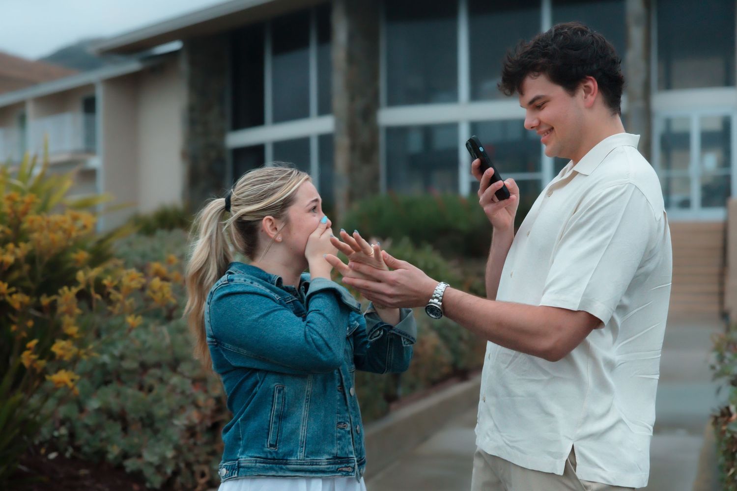 Man placing engagement ring on surprised woman's finger outside building during proposal.