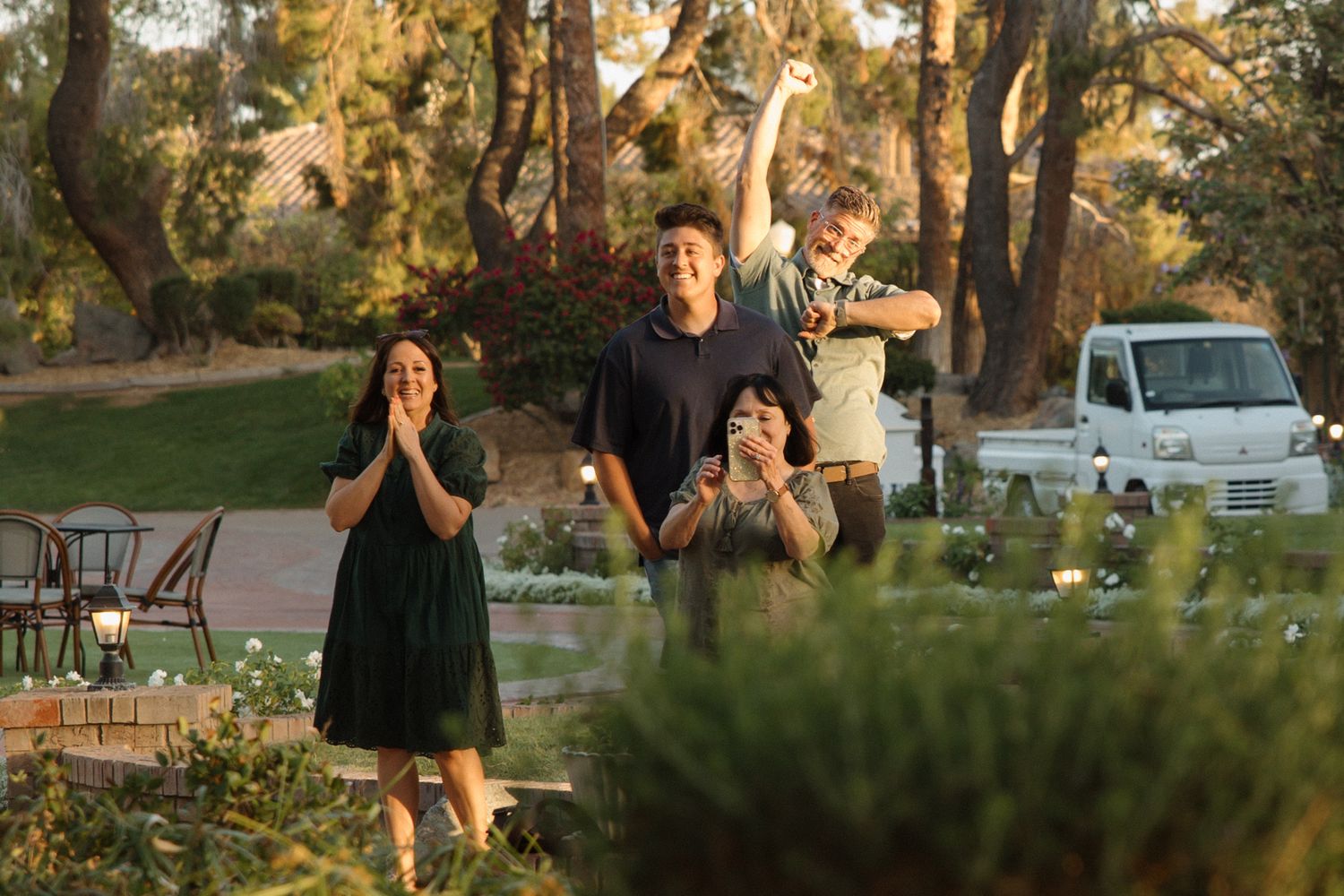 Family of four joyfully posing outdoors in a lush garden setting during golden hour, with trees and a white van in background.