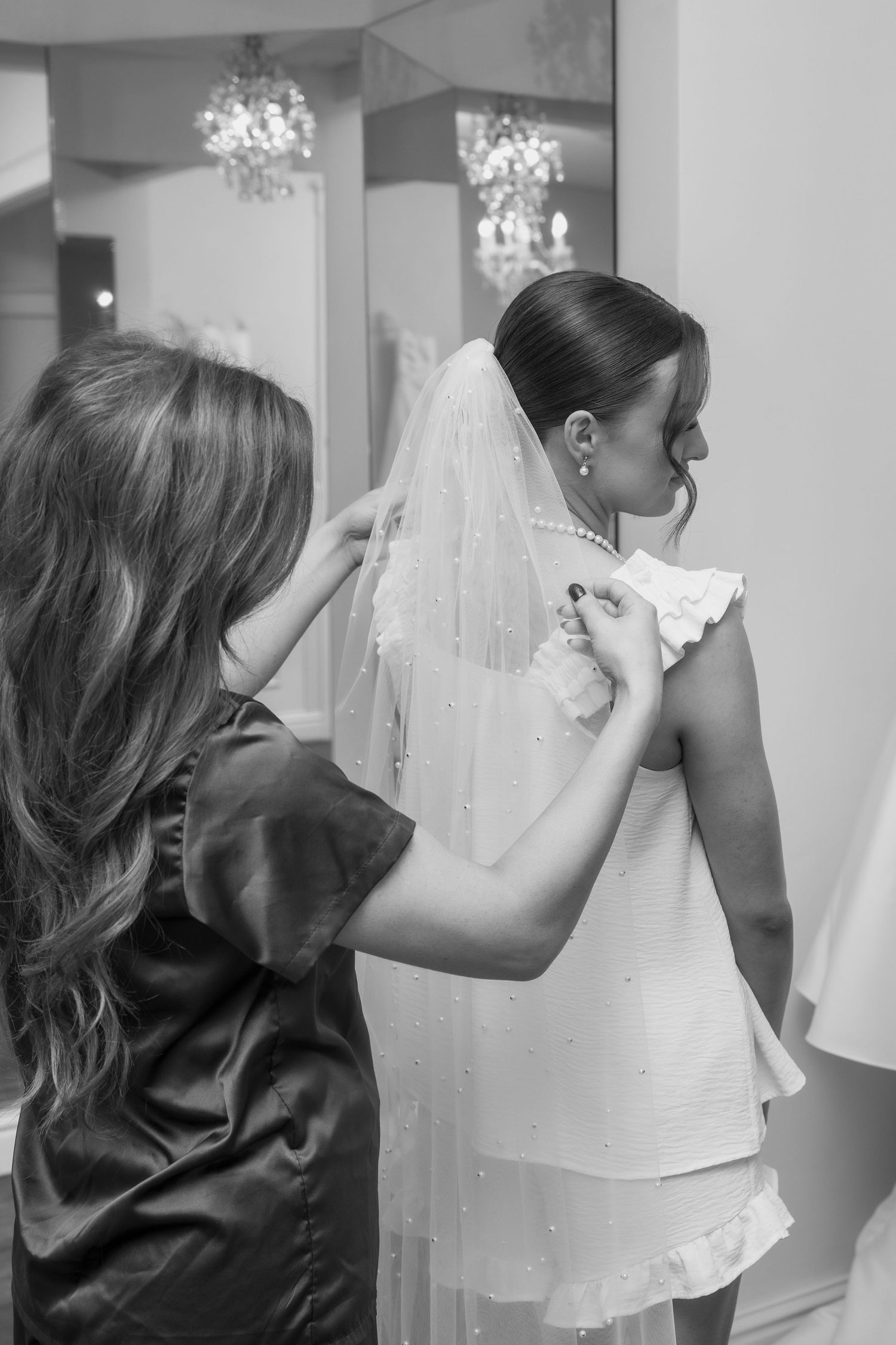 Black and white photo of bridesmaid helping bride with pearl-embellished veil during wedding preparations.