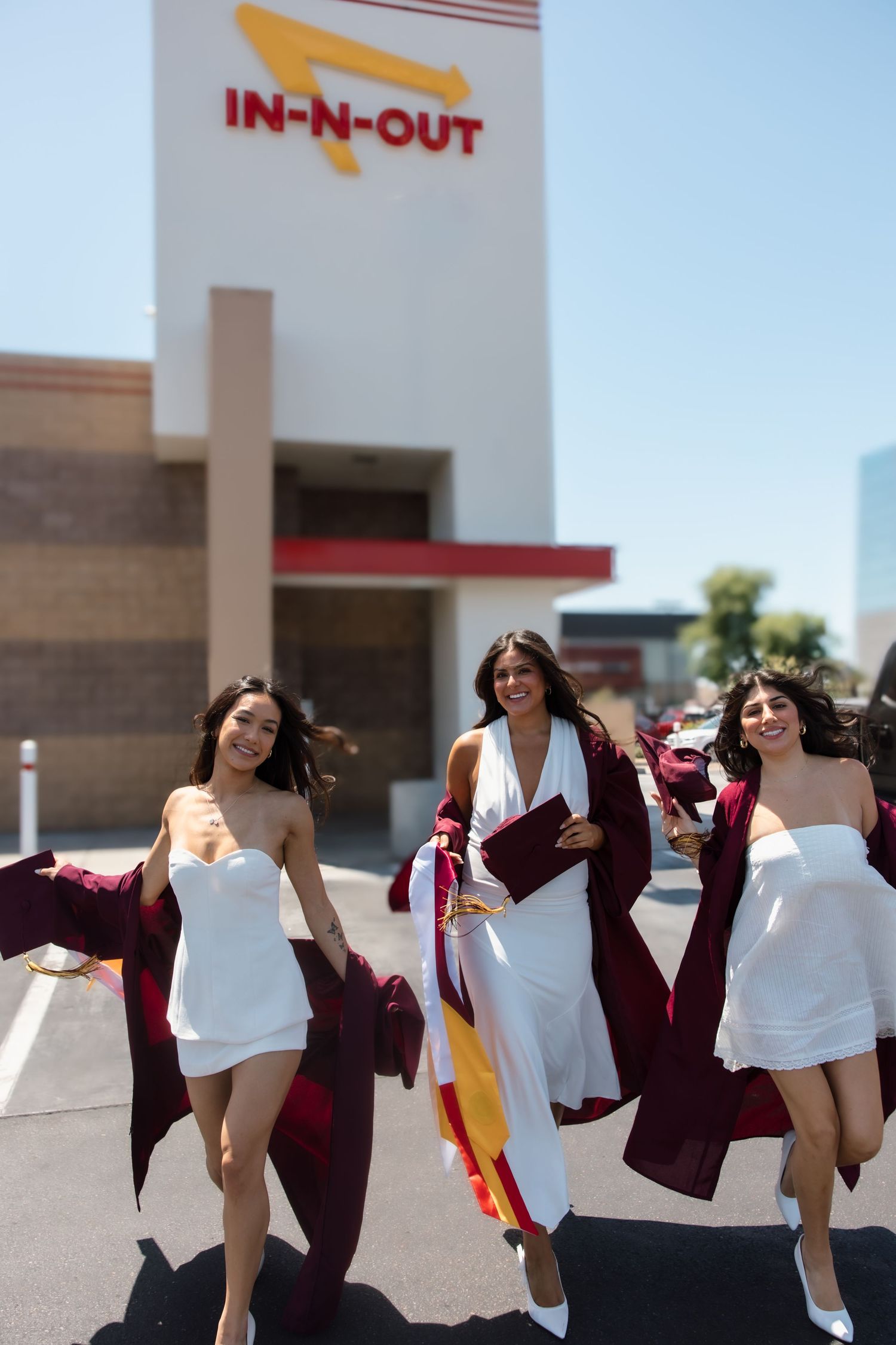 Three smiling graduates in white dresses and maroon gowns walking outside In-N-Out Burger.