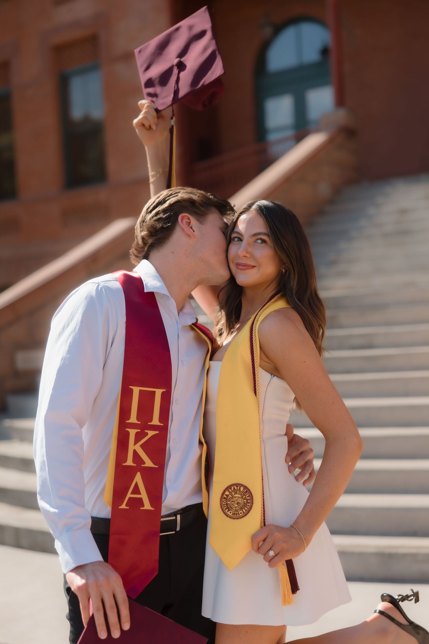 Graduate in Pi Kappa Alpha stole kissing woman in white dress holding maroon cap on campus steps.
