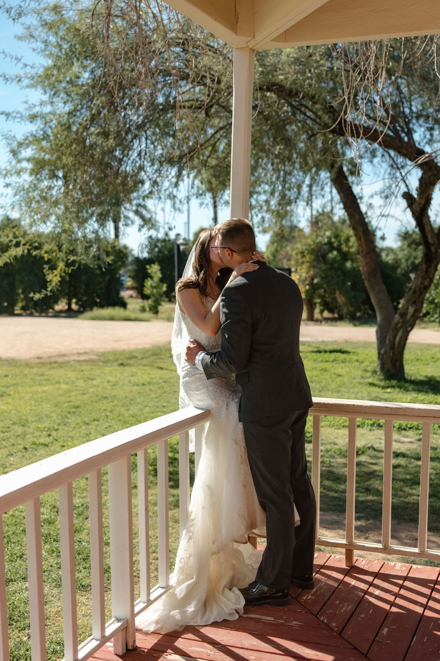 Bride and groom share a romantic kiss on a white wooden porch surrounded by lush green trees.