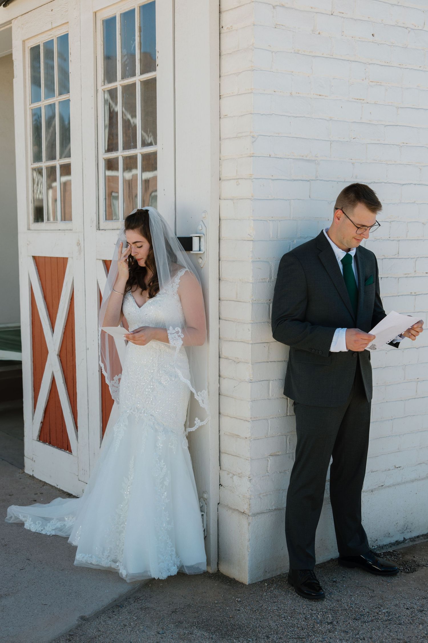 Bride in lace gown and groom in dark suit read vows separately outside white brick barn before wedding ceremony.