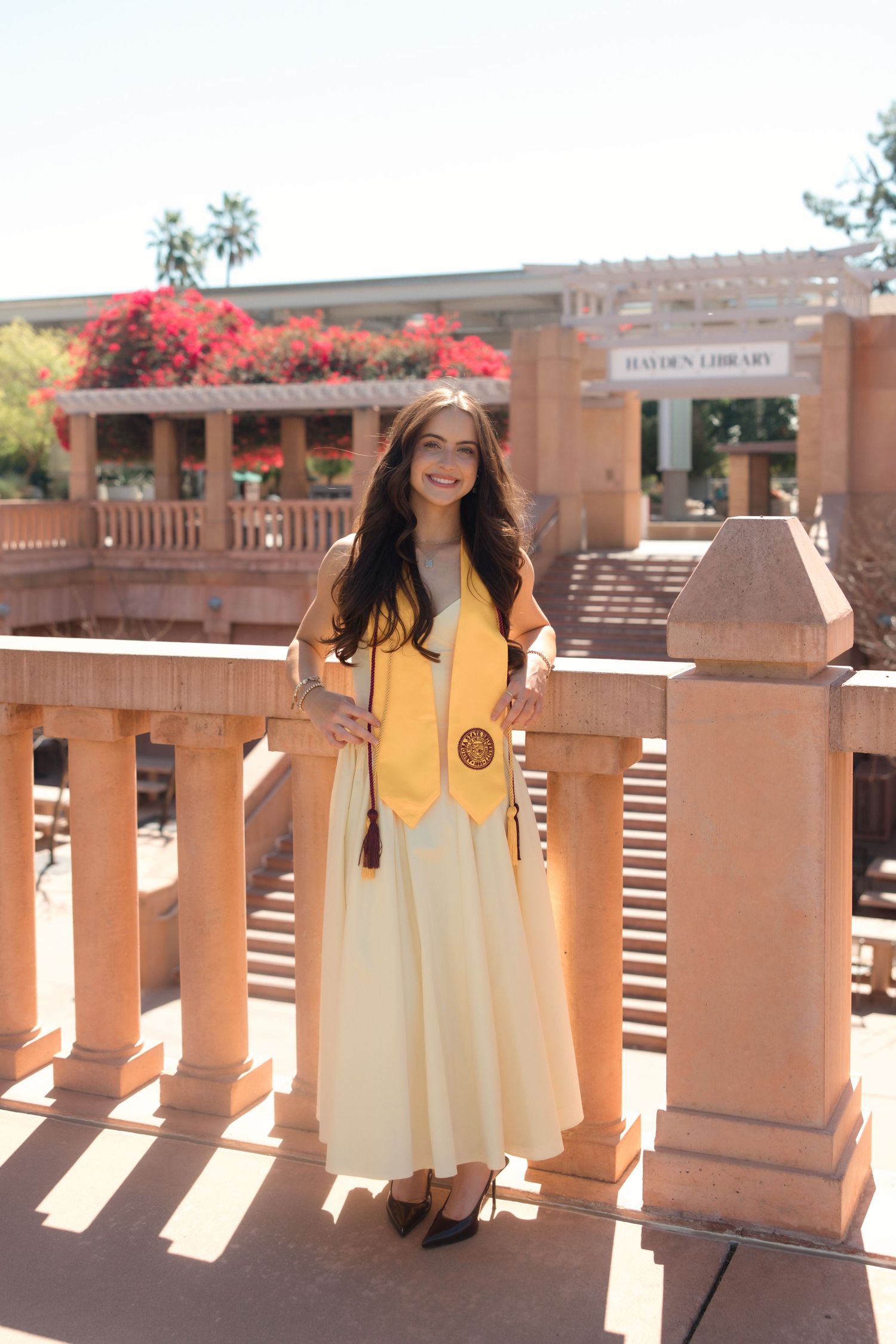 Young woman in graduation attire poses on a balcony outside Hayden Library at Arizona State University.