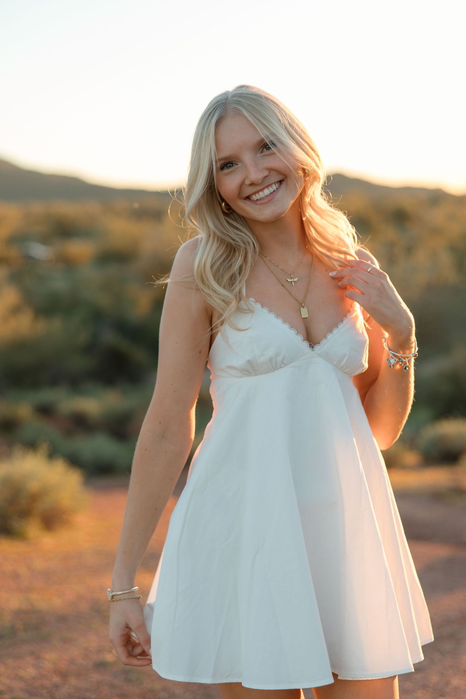 Joyful blonde woman in white spaghetti strap mini dress with gold jewelry smiling in desert sunset light.