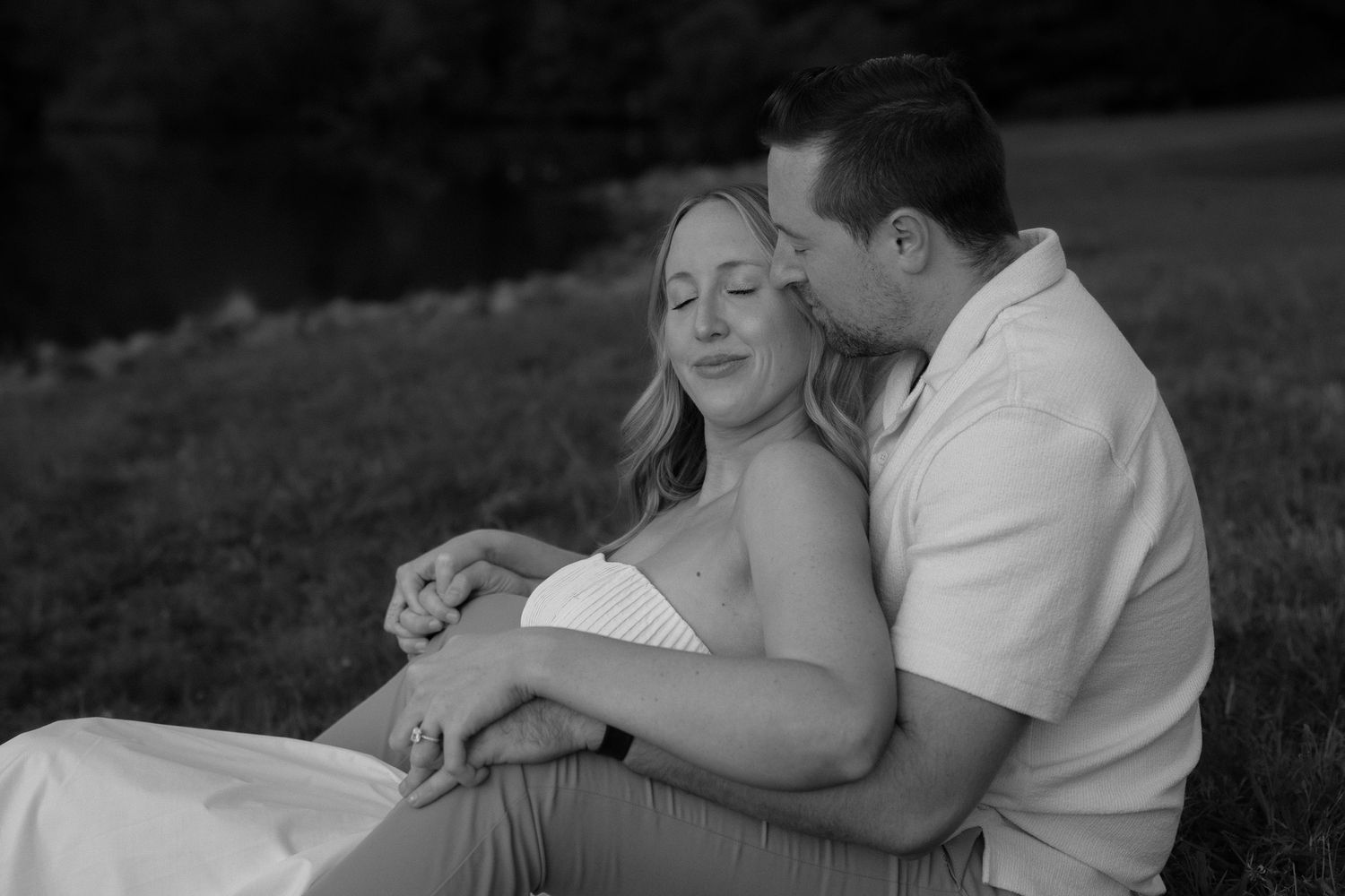 Black and white photo of a couple sitting on grass, woman leaning back into mans arms tenderly.