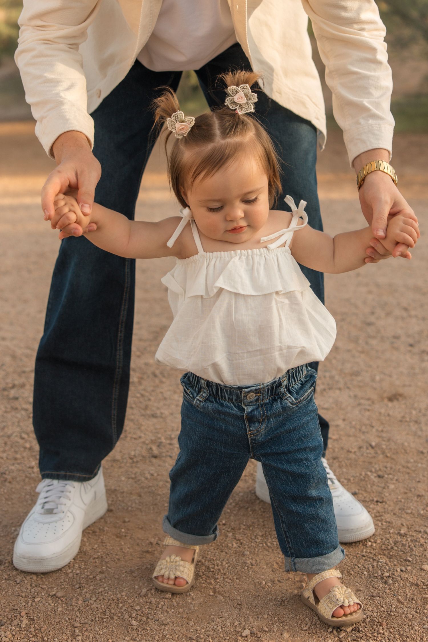 Toddler girl in white top and jeans taking first steps while adult holds her hands outdoors.
