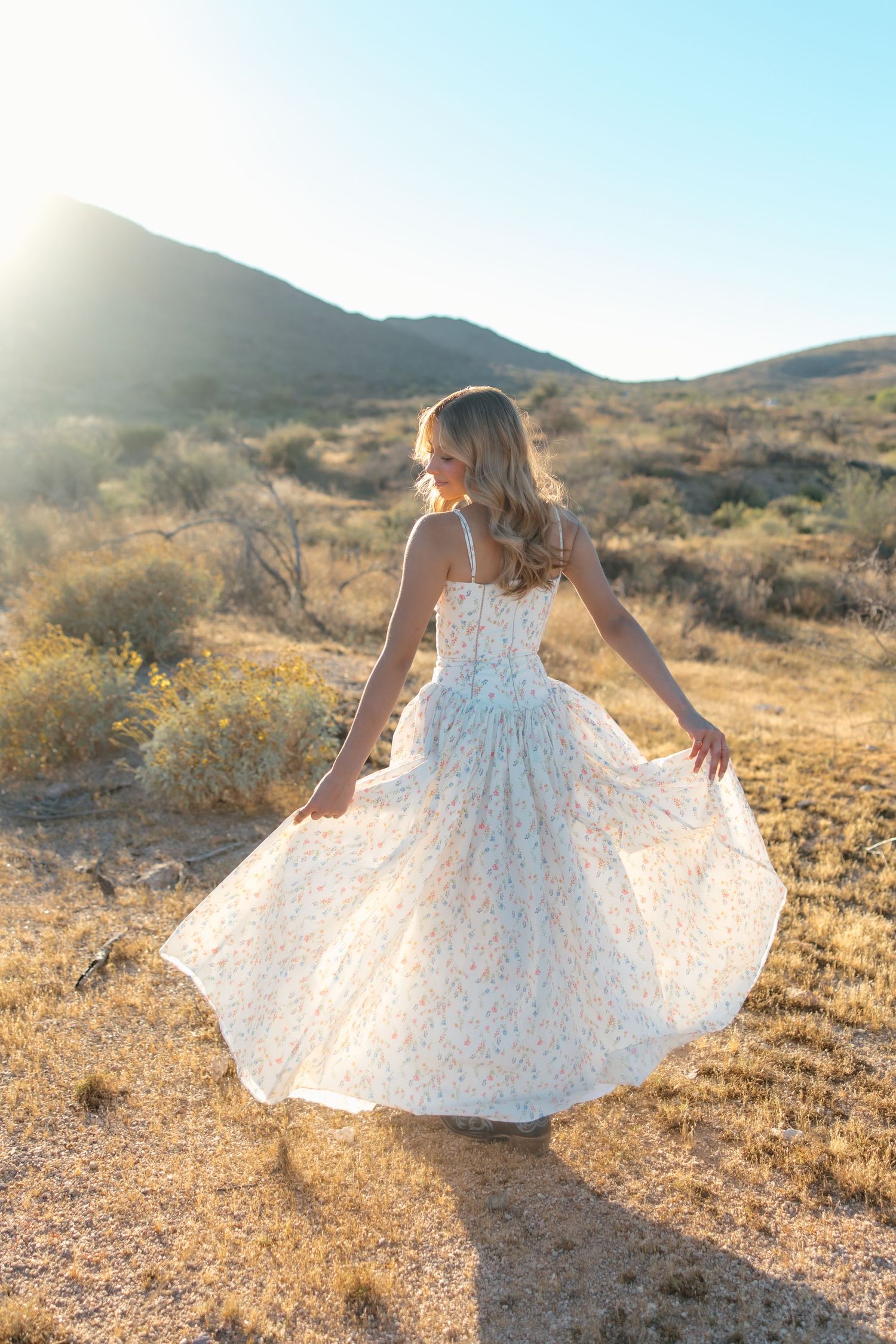 Woman twirling in a floral dress in a sunlit desert landscape with mountains in the background.