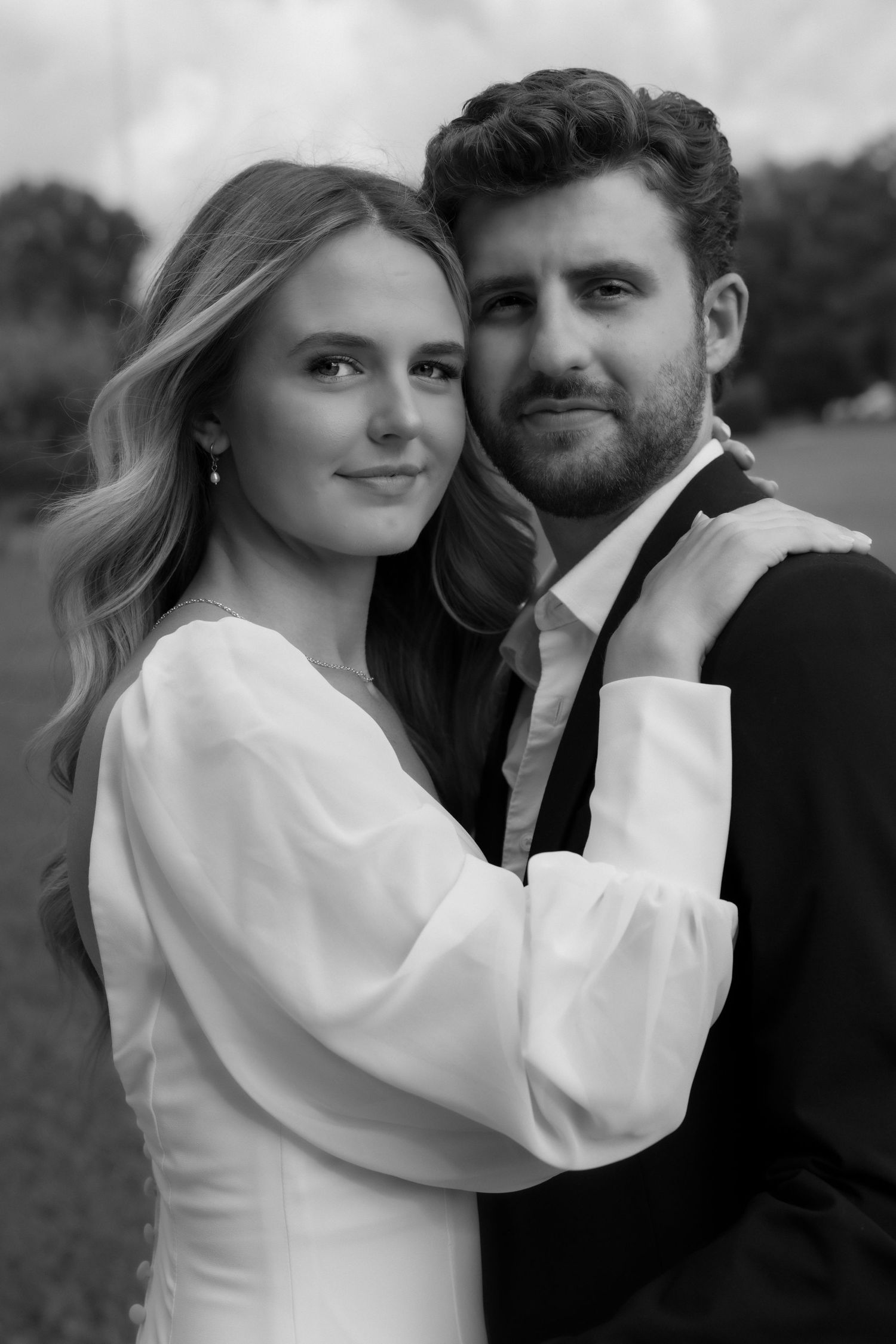 Black and white portrait of smiling bride in white dress embracing bearded groom in black suit outdoors.