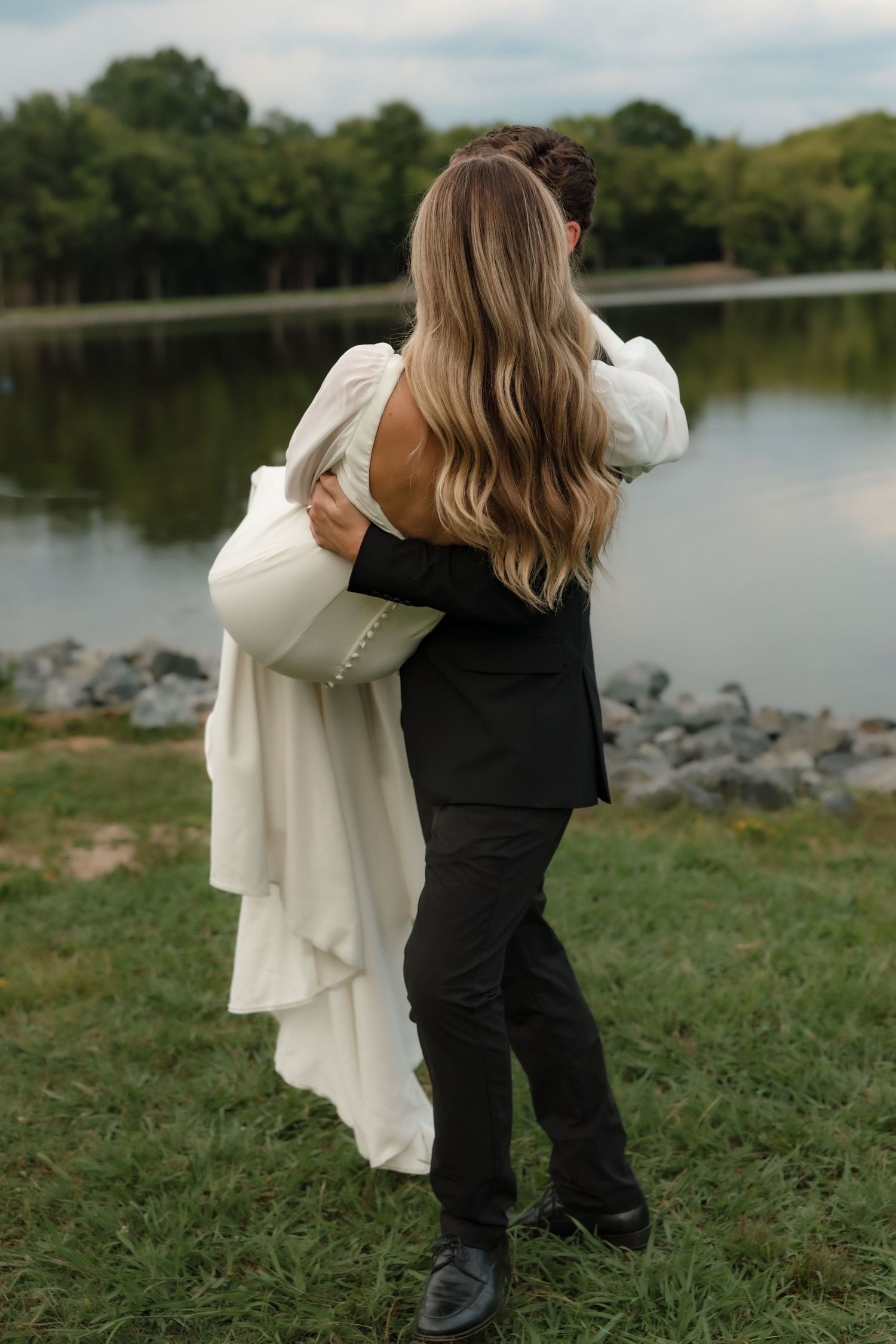 Groom lifting bride from behind near a peaceful lake, bride in white backless gown with long flowing hair.